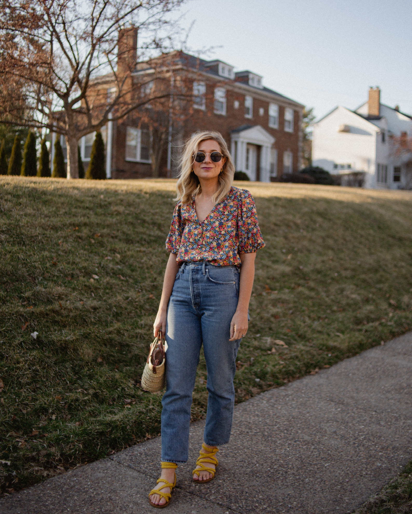 Karin Emily wears a floral top from Sezane, blue straight leg jeans from AGOLDE, and another pair of yellow wrap sandals from Sezane
