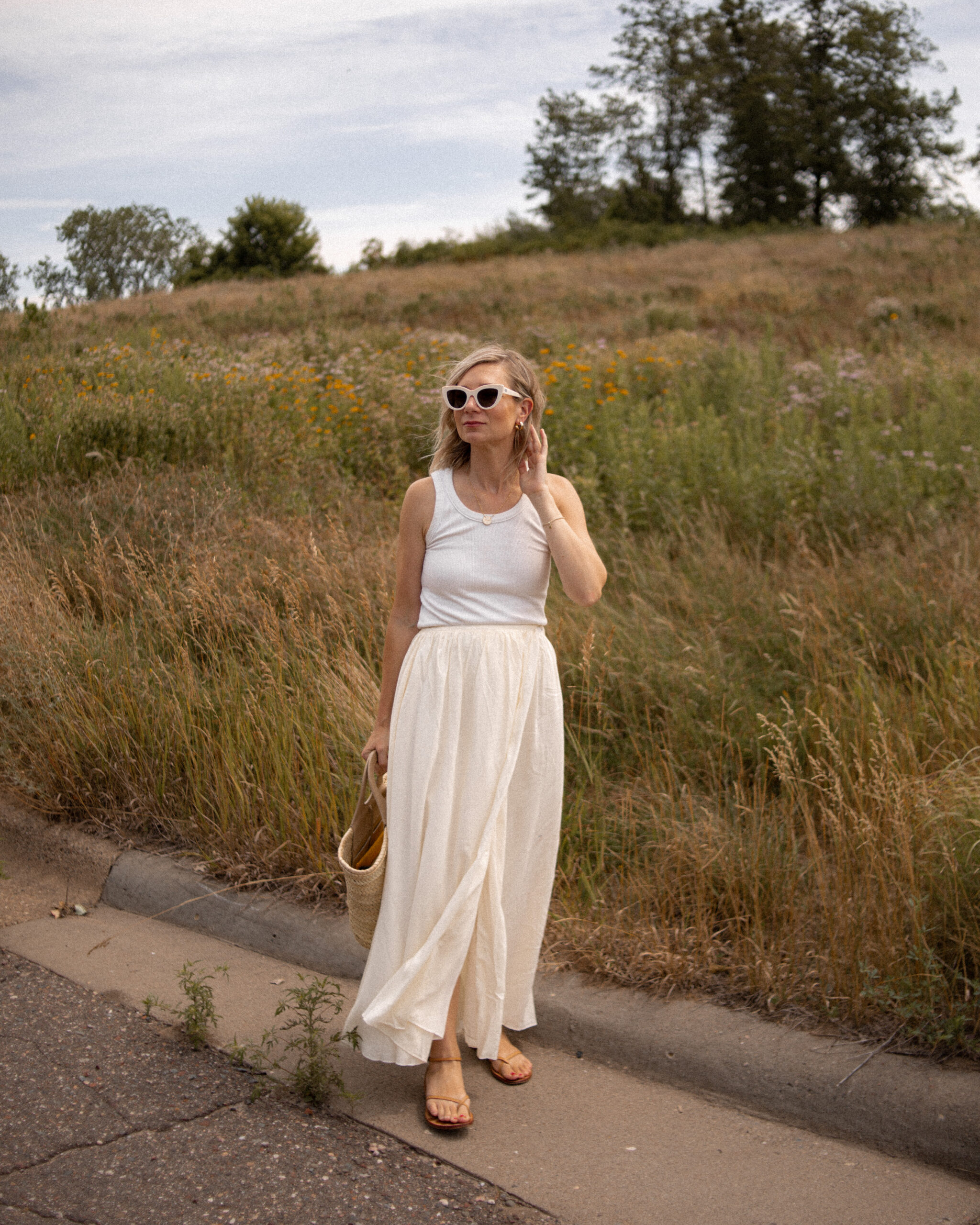 Karin Emily wears a cream wrap skirt and white tank top with white sunglasses and strappy sandals