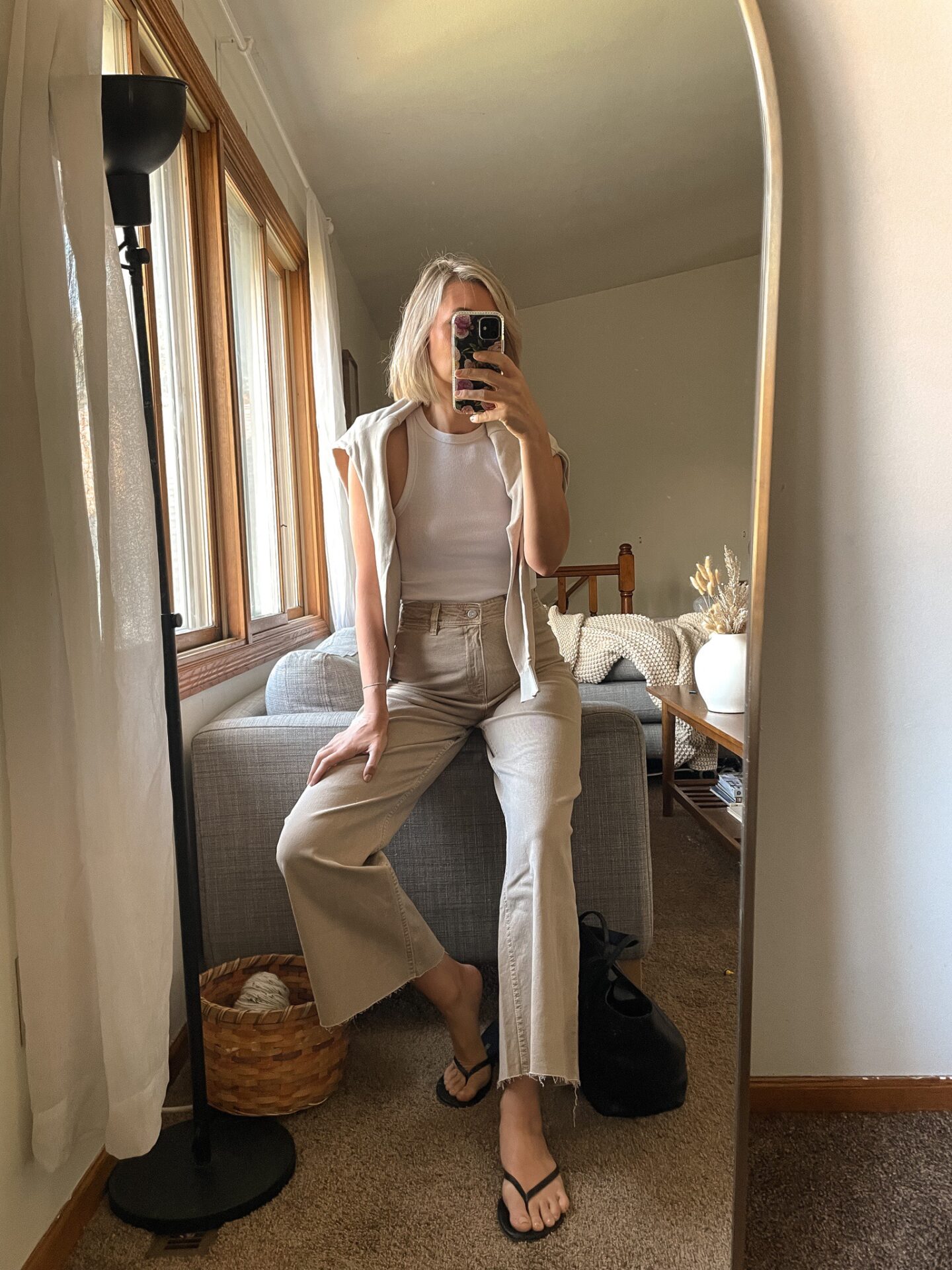 Karin Emily wears a khaki brown pair of wide leg jeans, a white anine bing tank, black flip flop sandals, and an oatmeal layering sweater over her shoulders