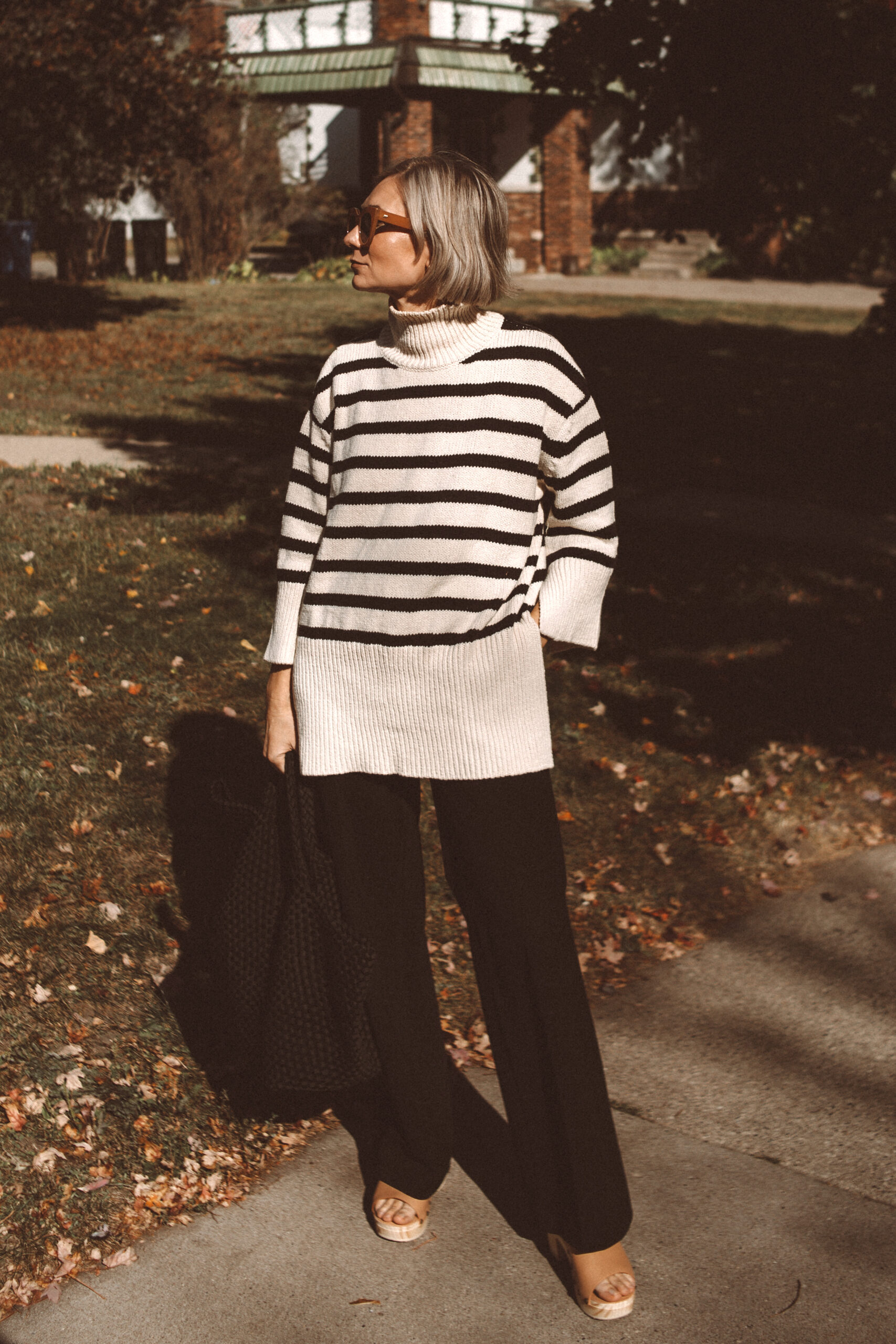 Karin Emily wears a pair of wooden blogs, striped turtleneck, black tote bag, and black wide leg pants