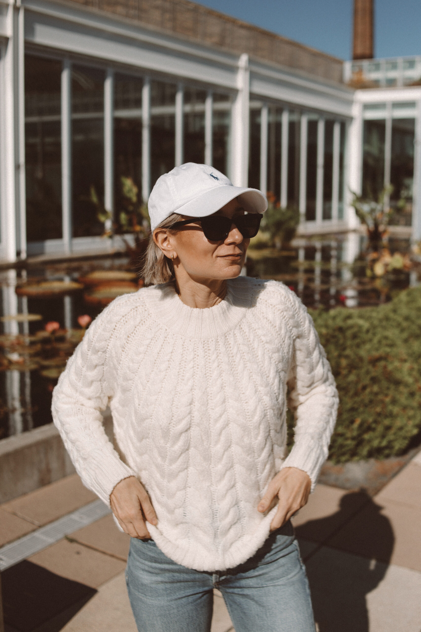 Karin Emily wears a cable knit sweater from Sezane with light wash straight leg jeans, cream sneakers, and a Ralph Lauren baseball hat