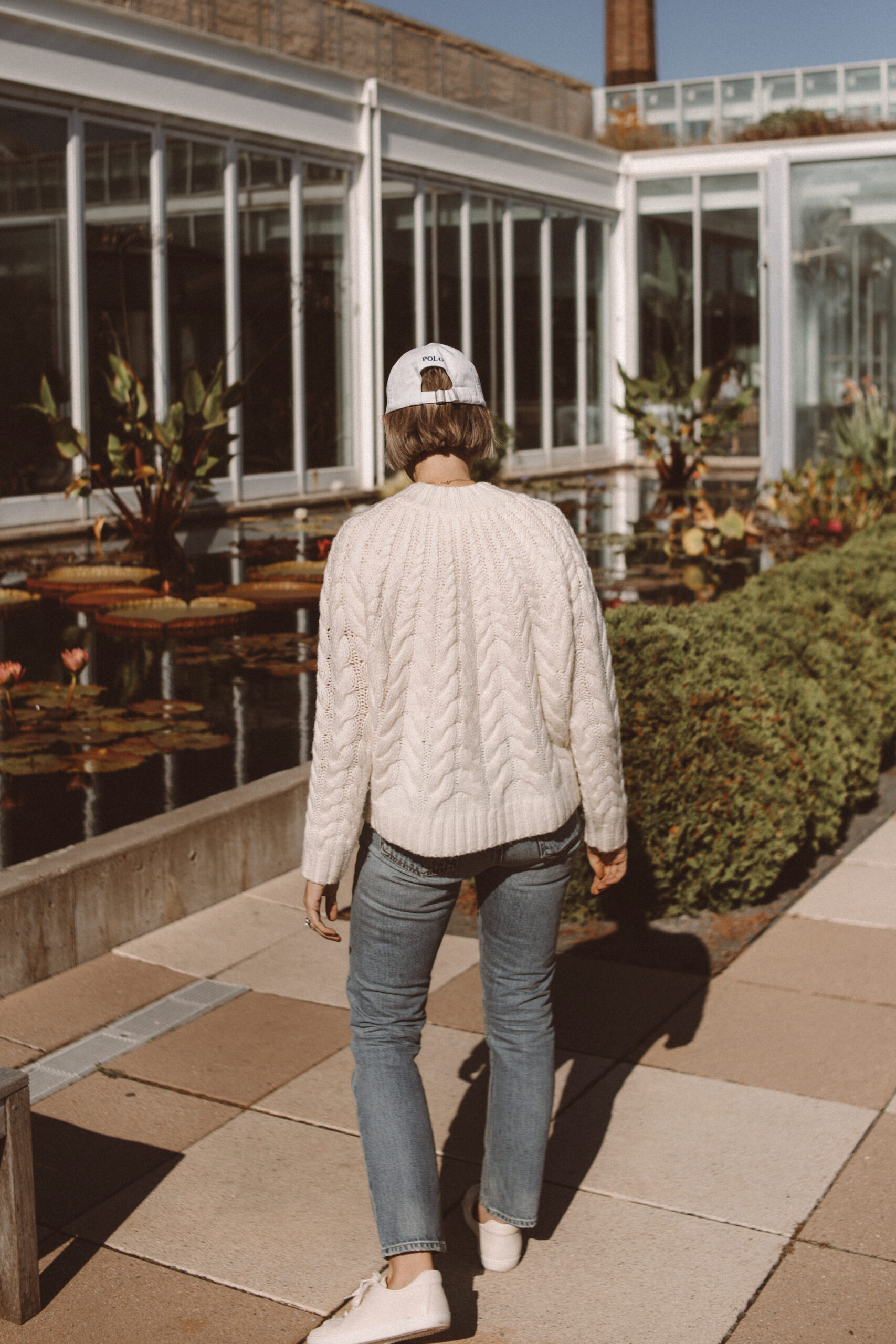 Karin Emily wears a cable knit sweater from Sezane with light wash straight leg jeans, cream sneakers, and a Ralph Lauren baseball hat