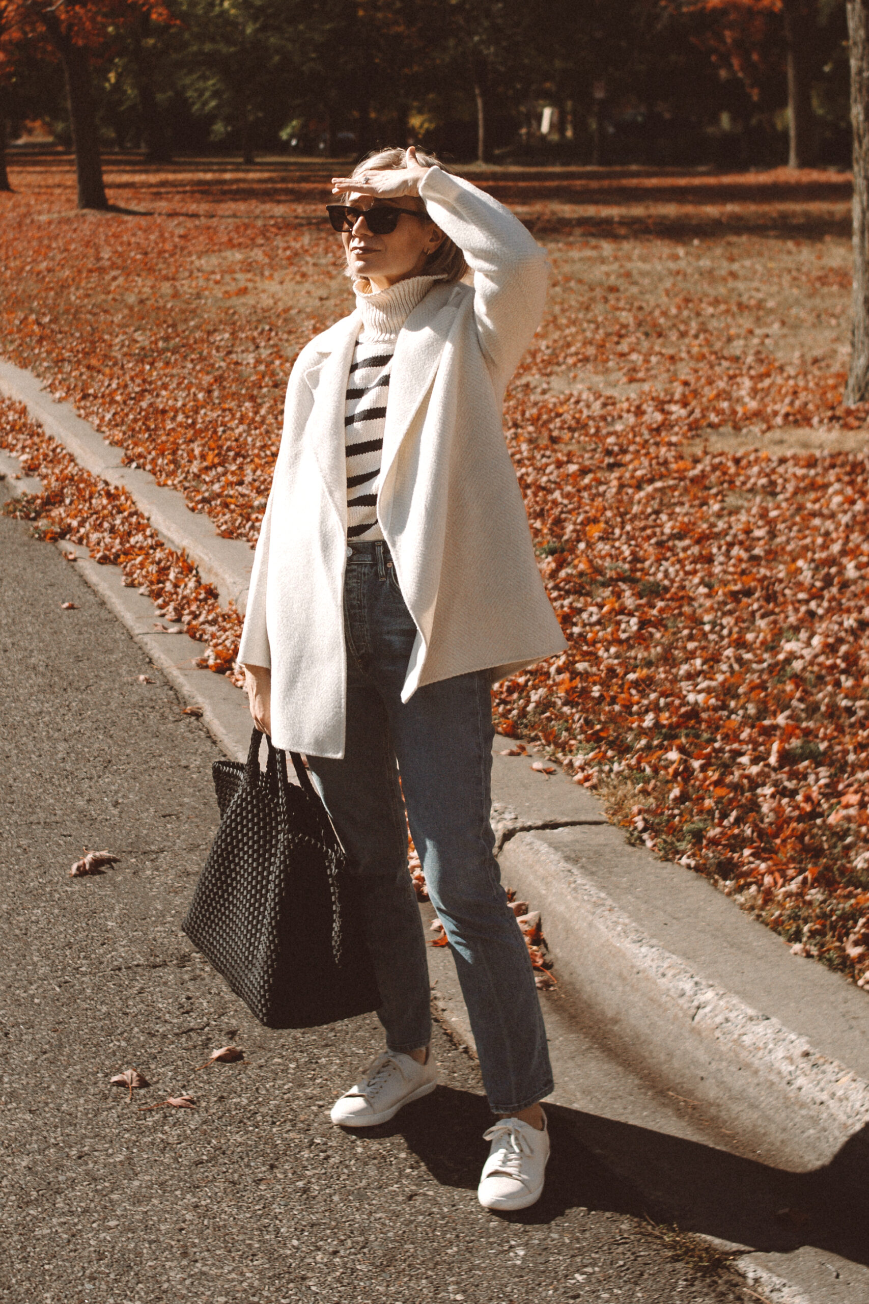 Karin Emily wears the Theory Clairene Jacket, a striped turtleneck sweater, citizens of humanity jolene jeans, sezane sneakers, and naghedi st barths large onyx bag while standing in front of a yard full of all leaves