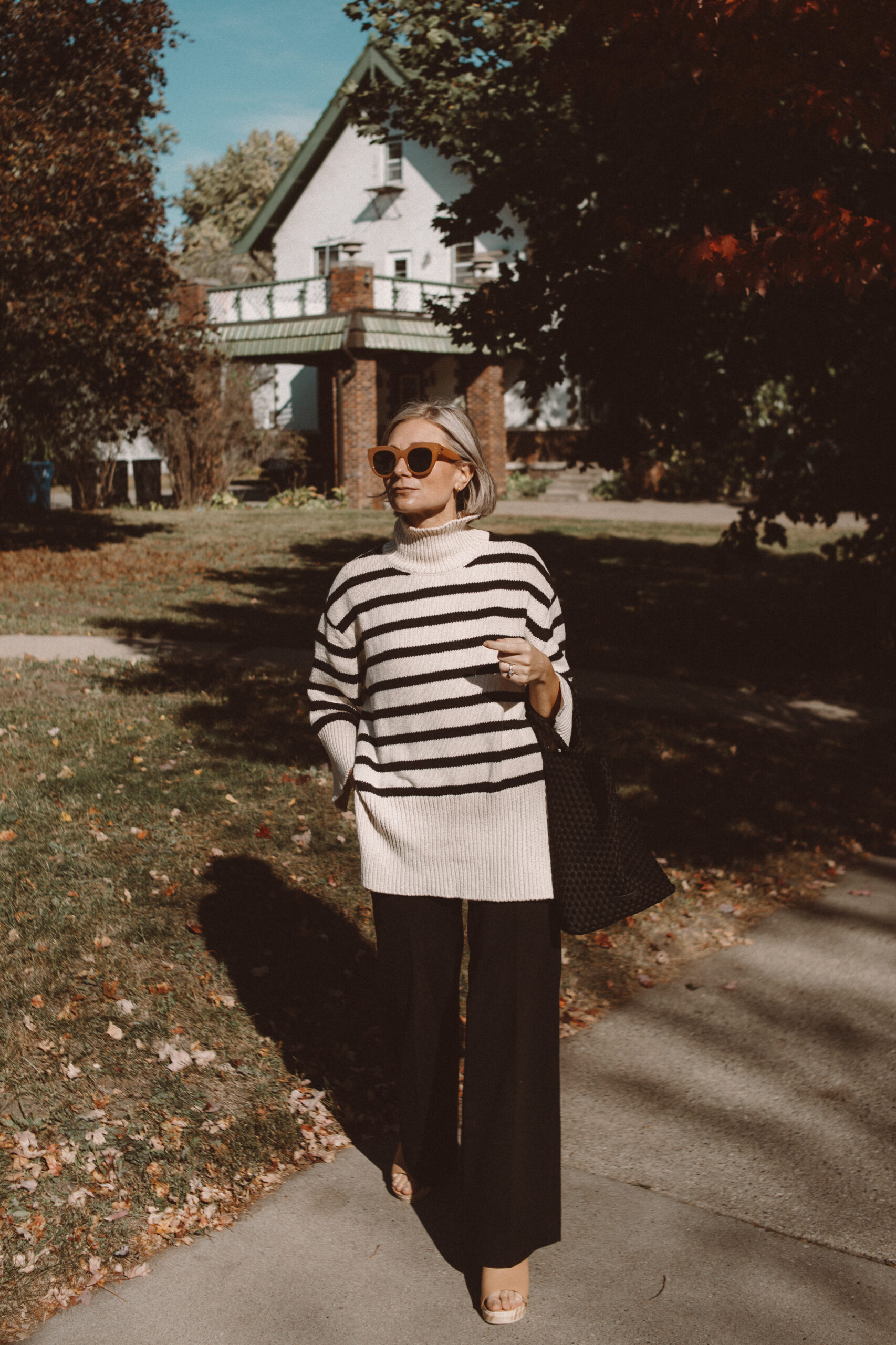 Karin Emily wears a pair of wooden blogs, striped turtleneck, black tote bag, and black wide leg pants