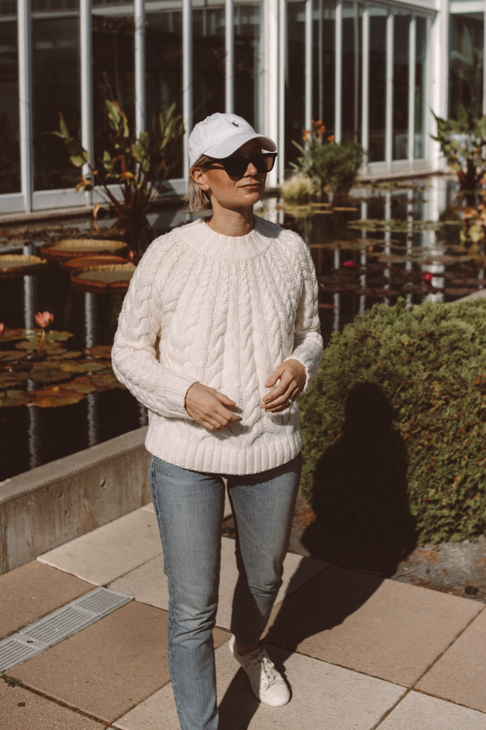 Karin Emily wears a cable knit sweater from Sezane with light wash straight leg jeans, cream sneakers, and a Ralph Lauren baseball hat