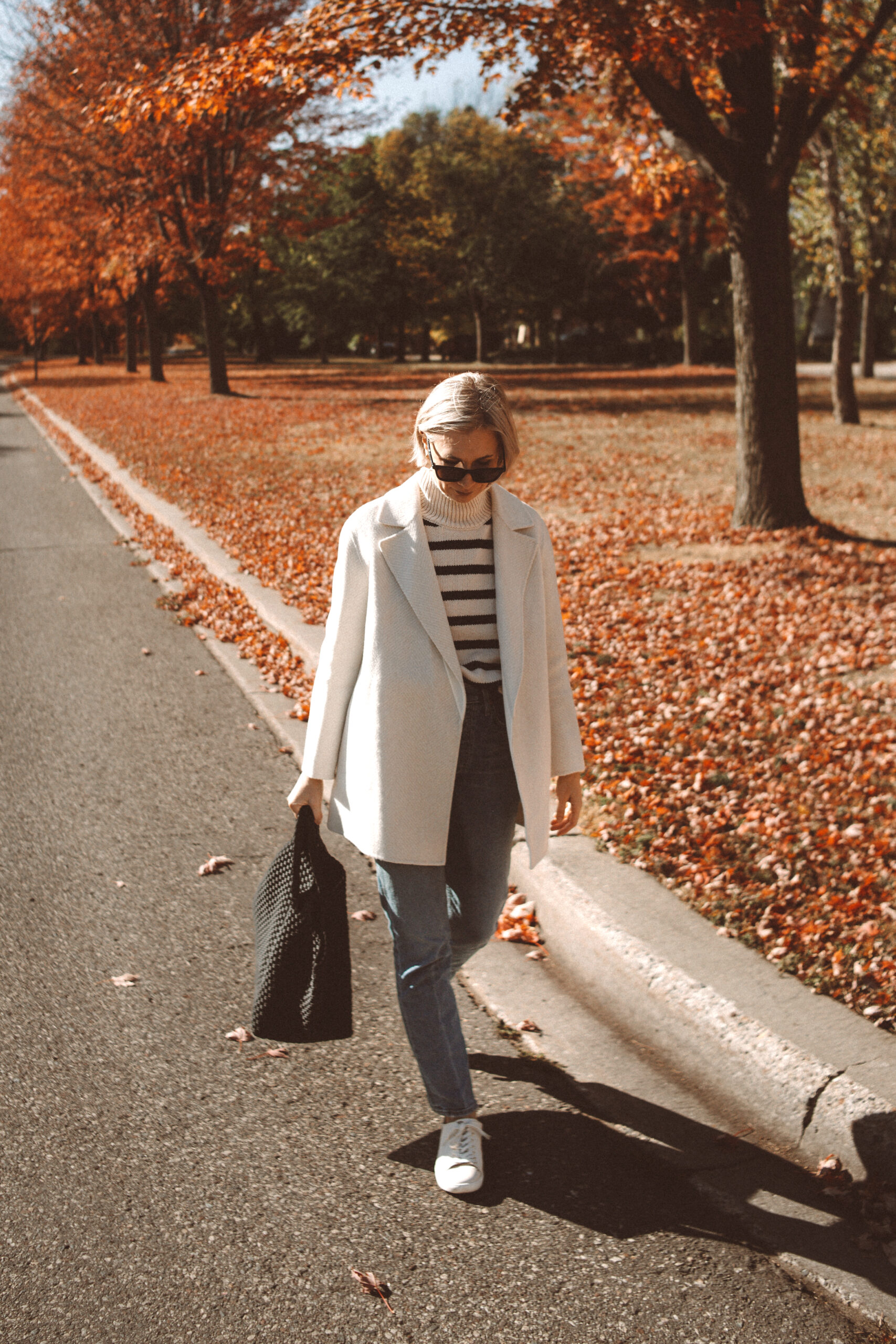 Karin Emily wears the Theory Clairene Jacket, a striped turtleneck sweater, citizens of humanity jolene jeans, sezane sneakers, and naghedi st barths large onyx bag while standing in front of a yard full of all leaves