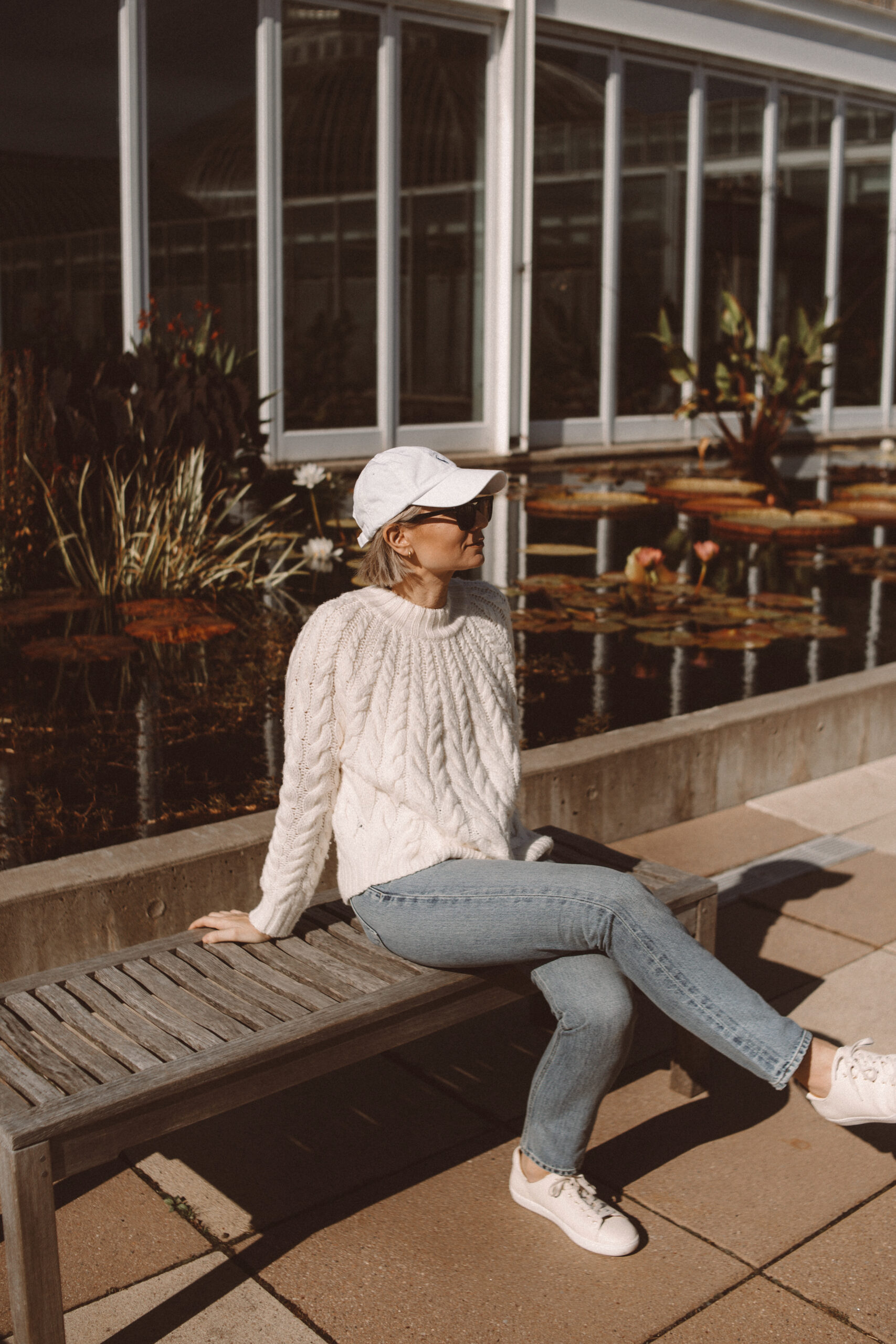 Karin Emily wears a cable knit sweater from Sezane with light wash straight leg jeans, cream sneakers, and a Ralph Lauren baseball hat