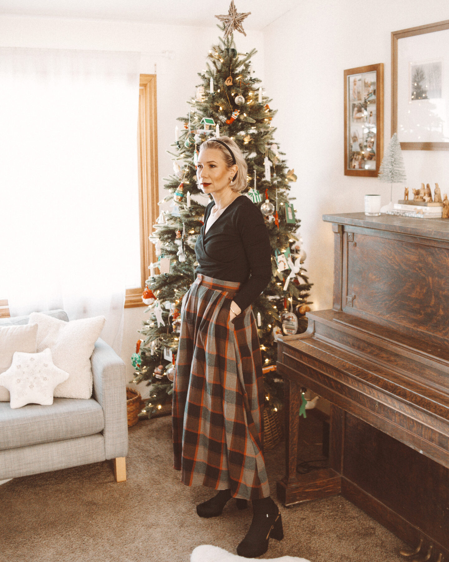 Karin Emily wears a holiday party outfit with a black wrap top, plaid maxi skirt, and black platform heels in front of her Christmas tree