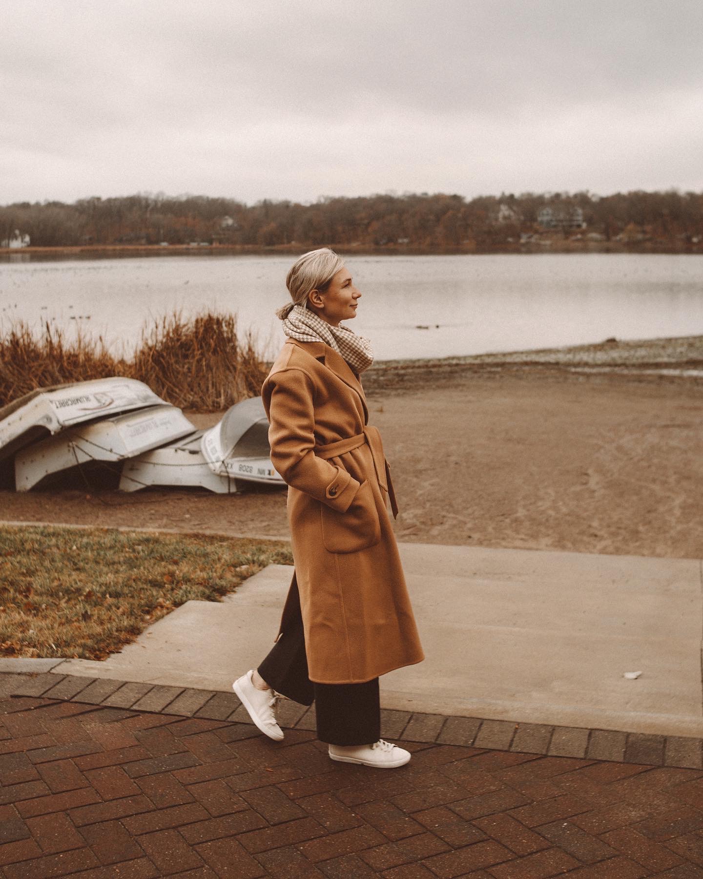 Karin Emily wears a houndstooth scarf, camel colored wool coat, black wide leg pants, cream colored sneakers