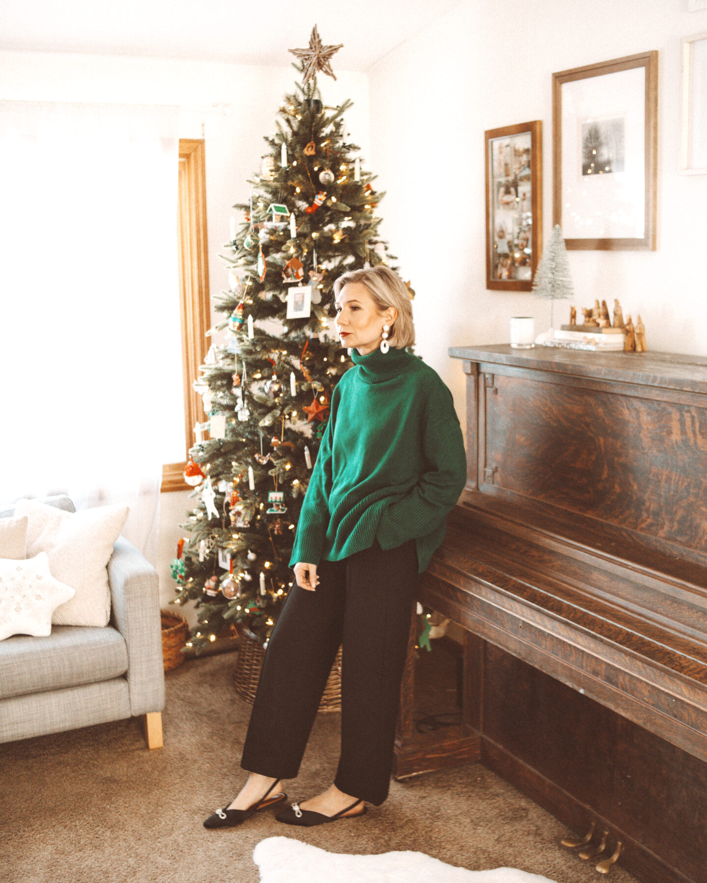 Karin Emily wears a holiday party outfit with a green sweater, black pants, and jeweled flats in front of her Christmas tree