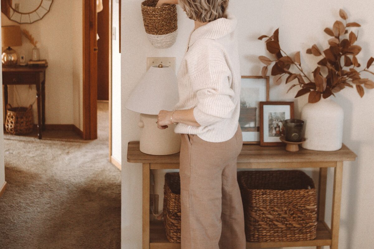 Karin Emily wears a white oversized sweater over tan linen pants with slippers in front of a console table