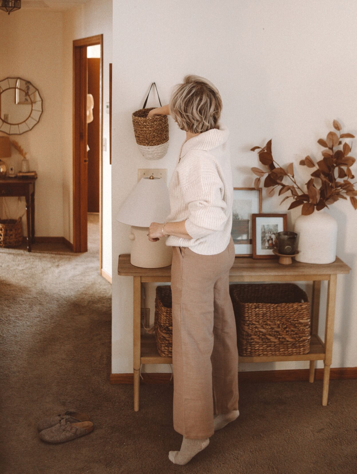 Karin Emily wears a white oversized sweater over tan linen pants with slippers in front of a console table