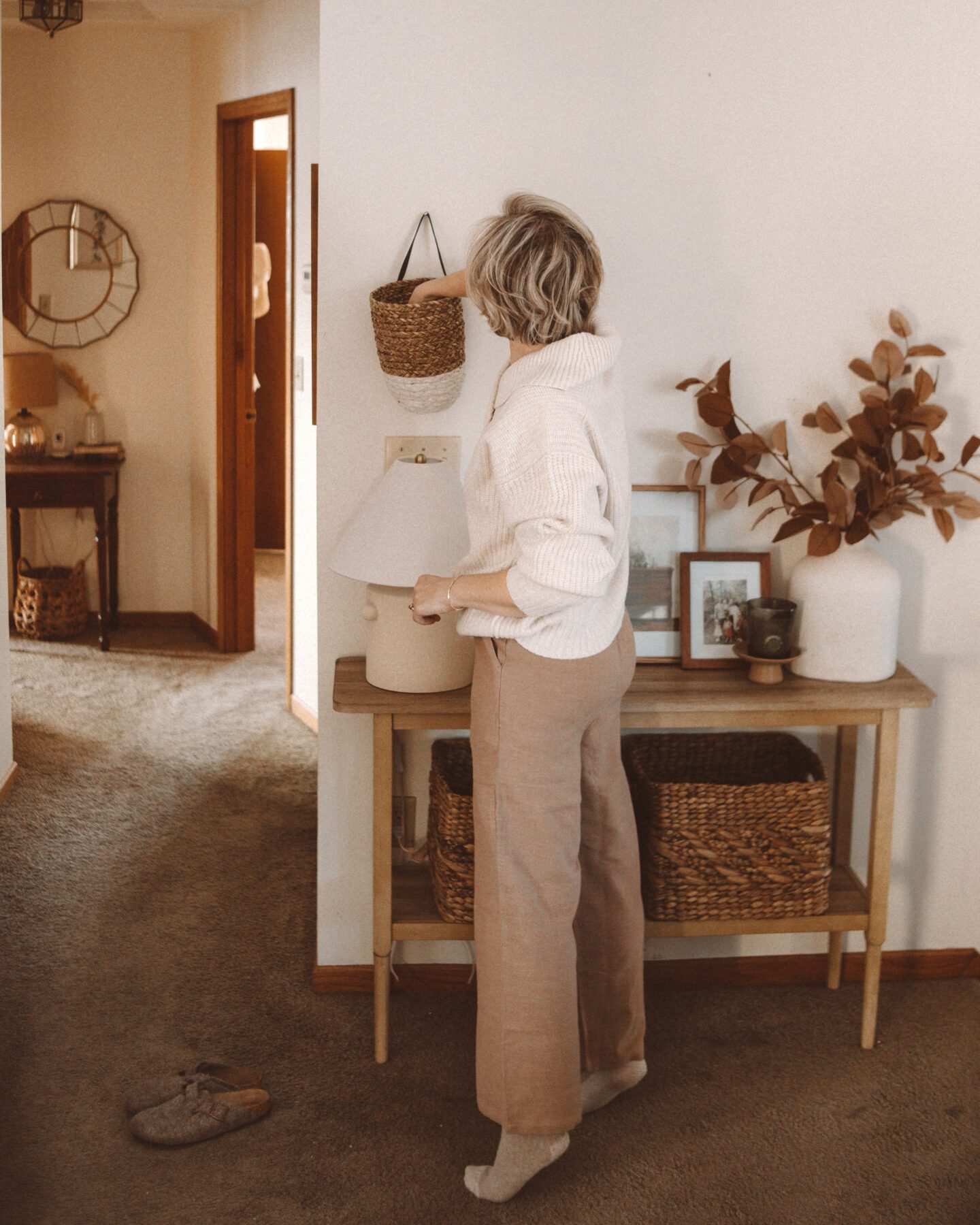 Karin Emily wears a white oversized sweater over tan linen pants with slippers in front of a console table 