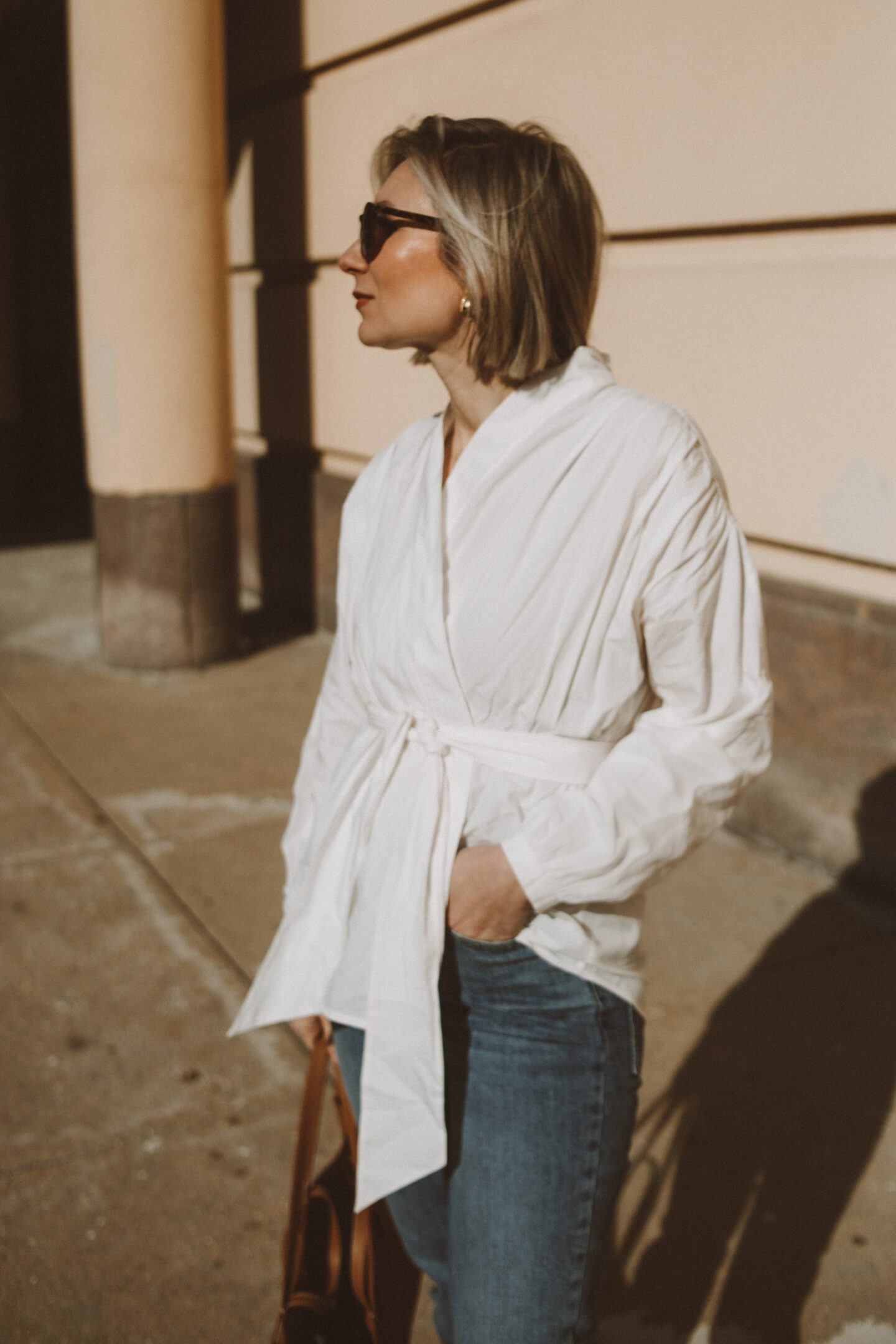 Karin Emily wears a white cotton wrap blouse with a pair of classic tortoiseshell sunglasses nd blue jeans