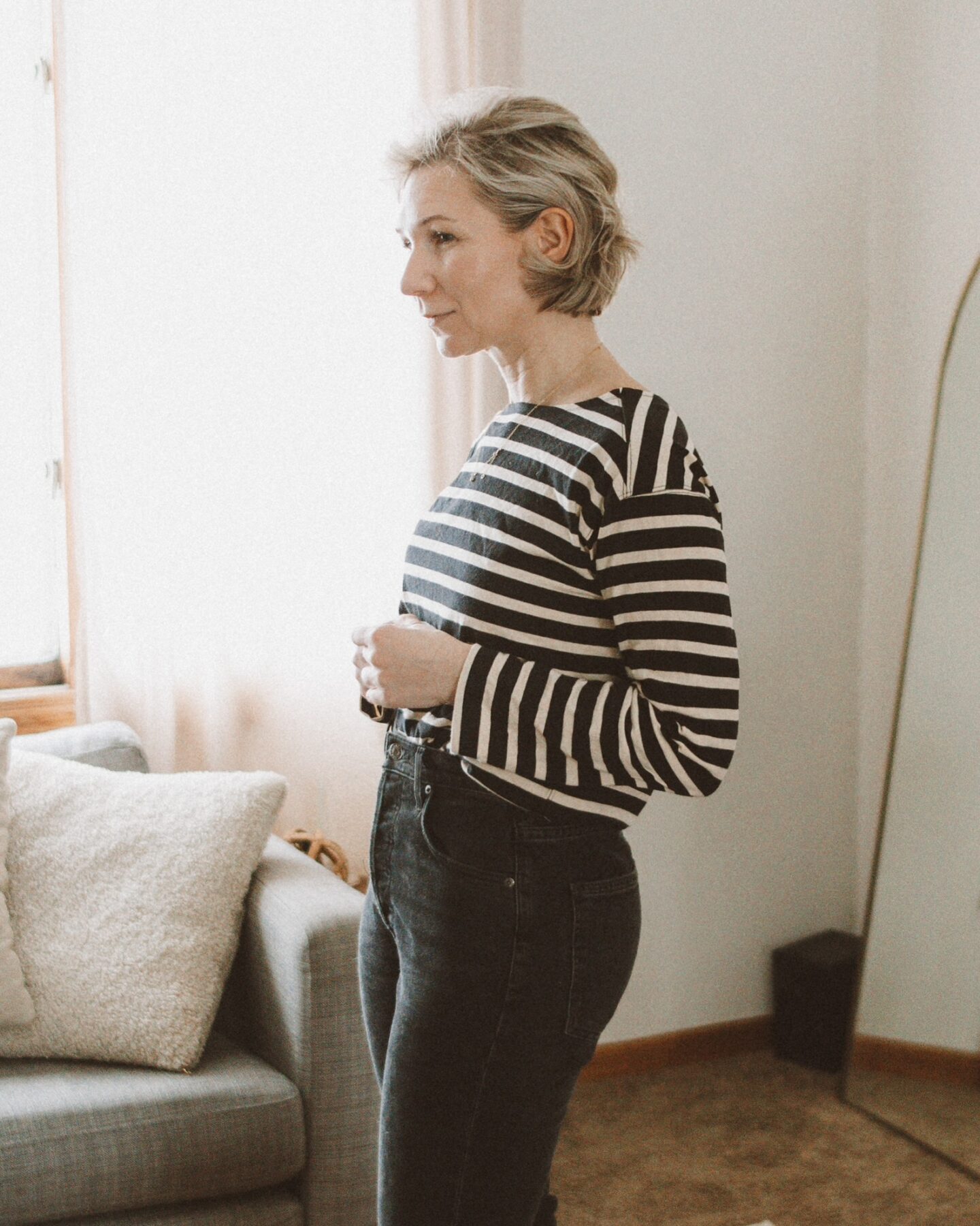 Karin Emily stands in front of her living room couch and standing mirror in her Agolde Freya jeans and Everlane breton stripe tee