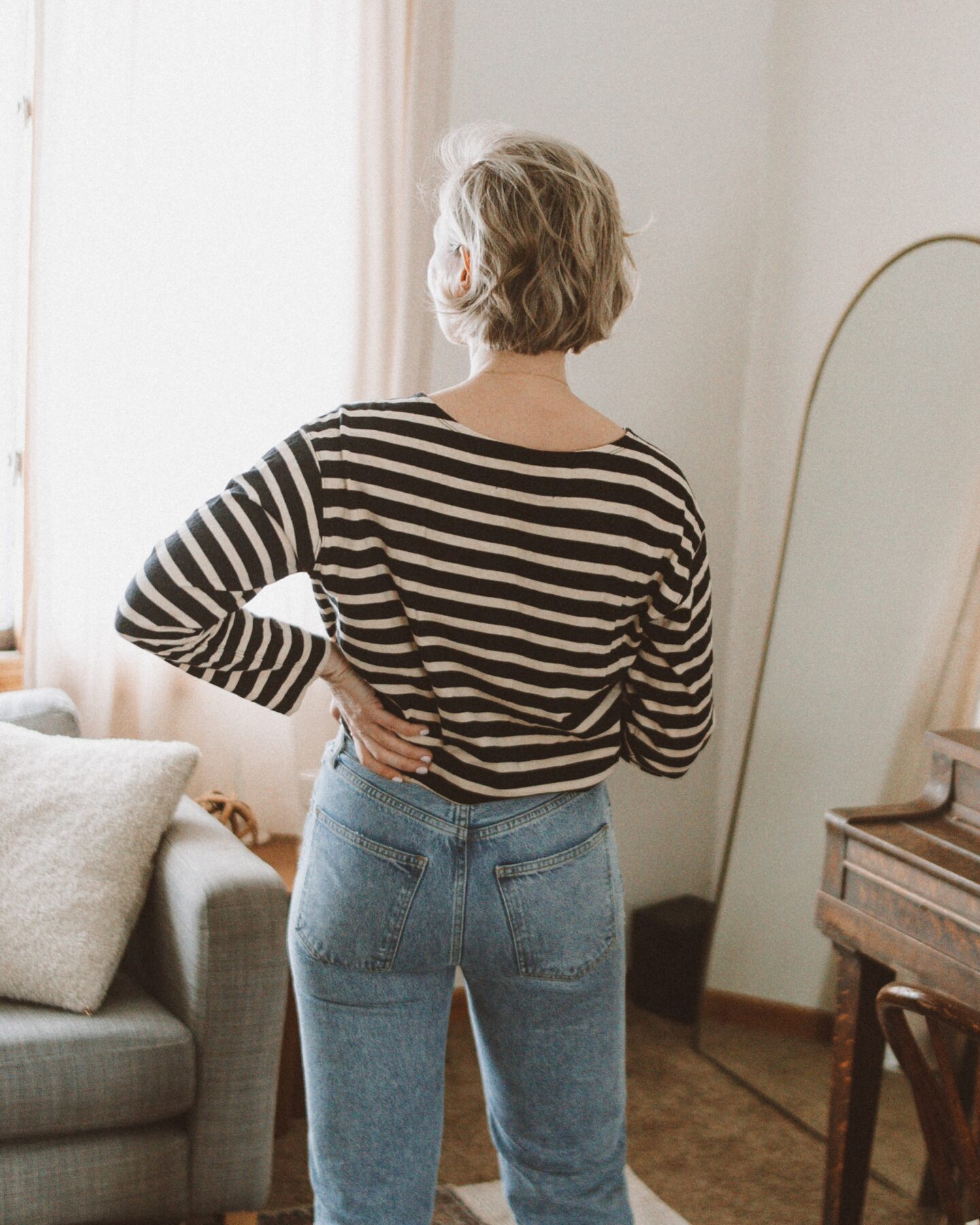 Karin Emily stands in front of her living room couch and standing mirror in her Agolde Lana jeans and Everlane breton stripe tee