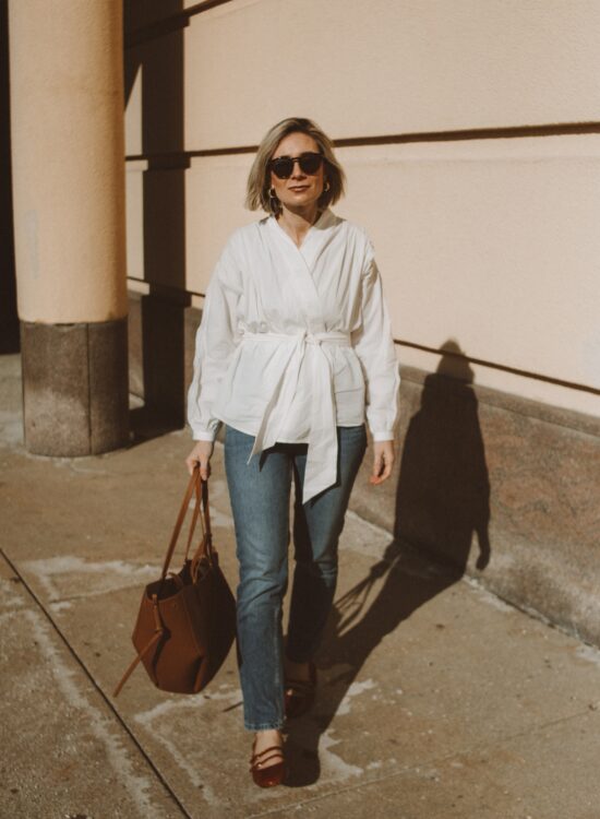 Karin Emily wears a white cotton wrap blouse with a pair of classic tortoiseshell sunglasses, straight leg blue jeans, a brown tote bag from Polene and brown patent leather slingback heels from Sezane