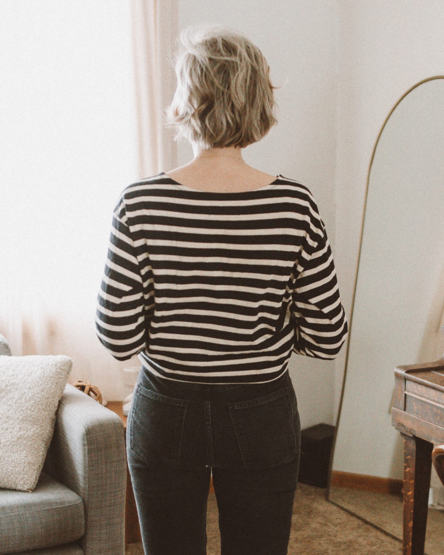 Karin Emily stands in front of her living room couch and standing mirror in her Agolde Freya jeans and Everlane breton stripe tee