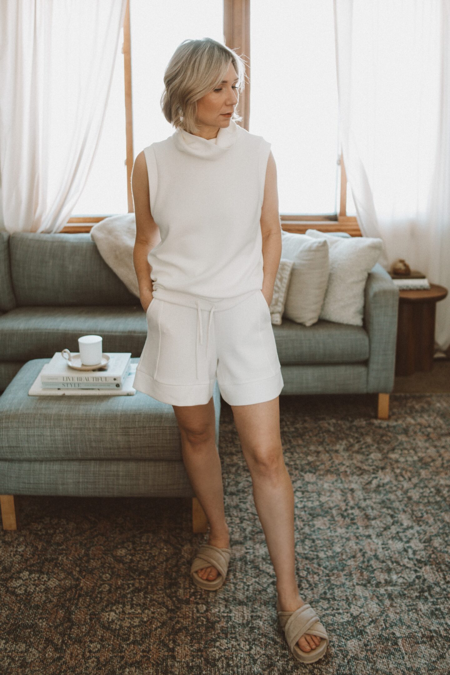 Karin Emily stands on her rug in front of her gray sofa and ottoman in her living room wearing a white top and shorts from Varley