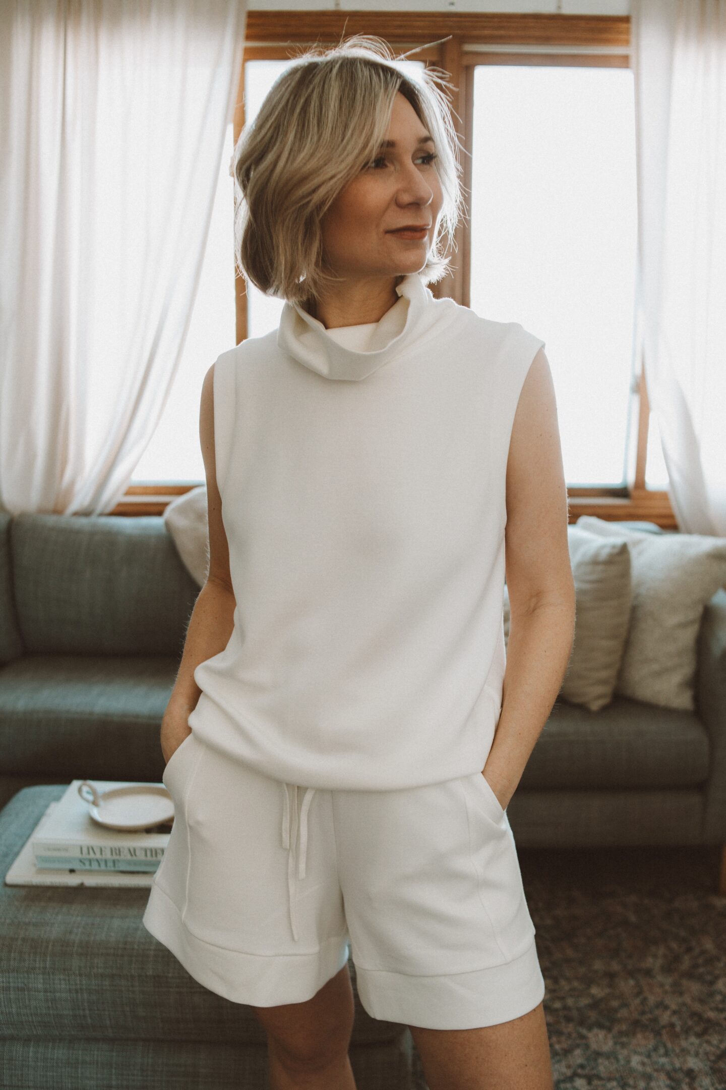 Karin Emily stands on her rug in front of her gray sofa and ottoman in her living room wearing a white top and shorts from Varley and shares the best style for spring from Varley
