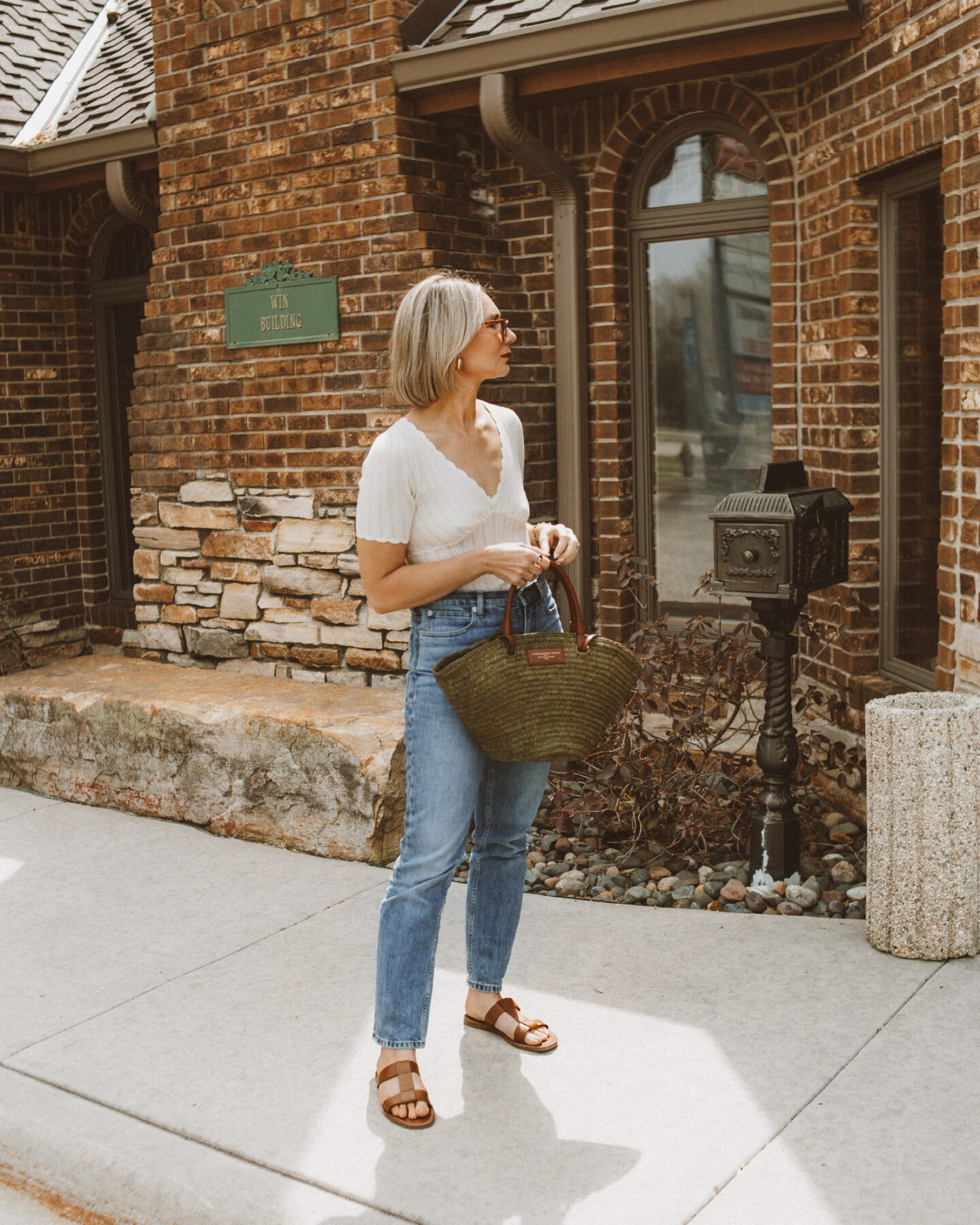 Karin Emily stands in front of a brick building wearing a white v neck tee from Sezane, a pair of blue jeans, and a khaki green justine basket bag