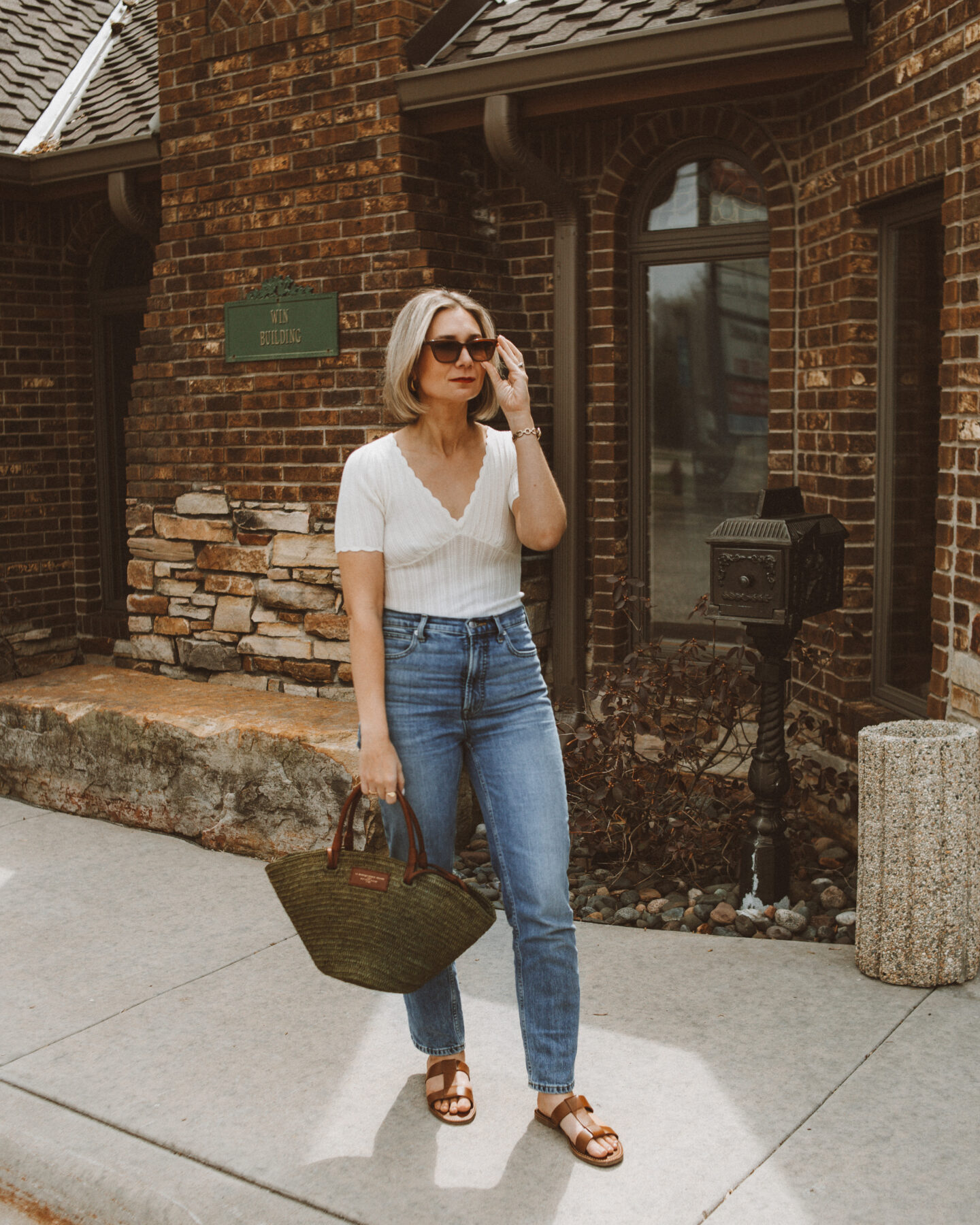 Karin Emily stands in front of a brick building wearing a white v neck tee from Sezane, a pair of blue jeans, and a khaki green justine basket bag