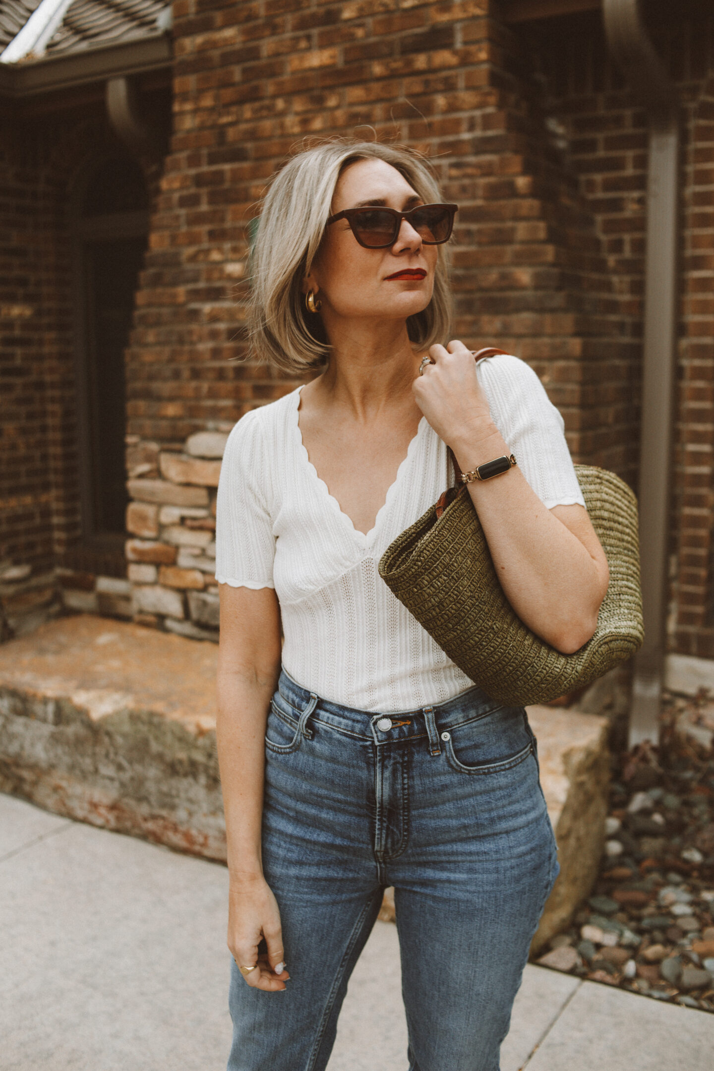 Karin Emily stands in front of a brick building wearing a white v neck tee from Sezane, a pair of blue jeans, and a khaki green justine basket bag