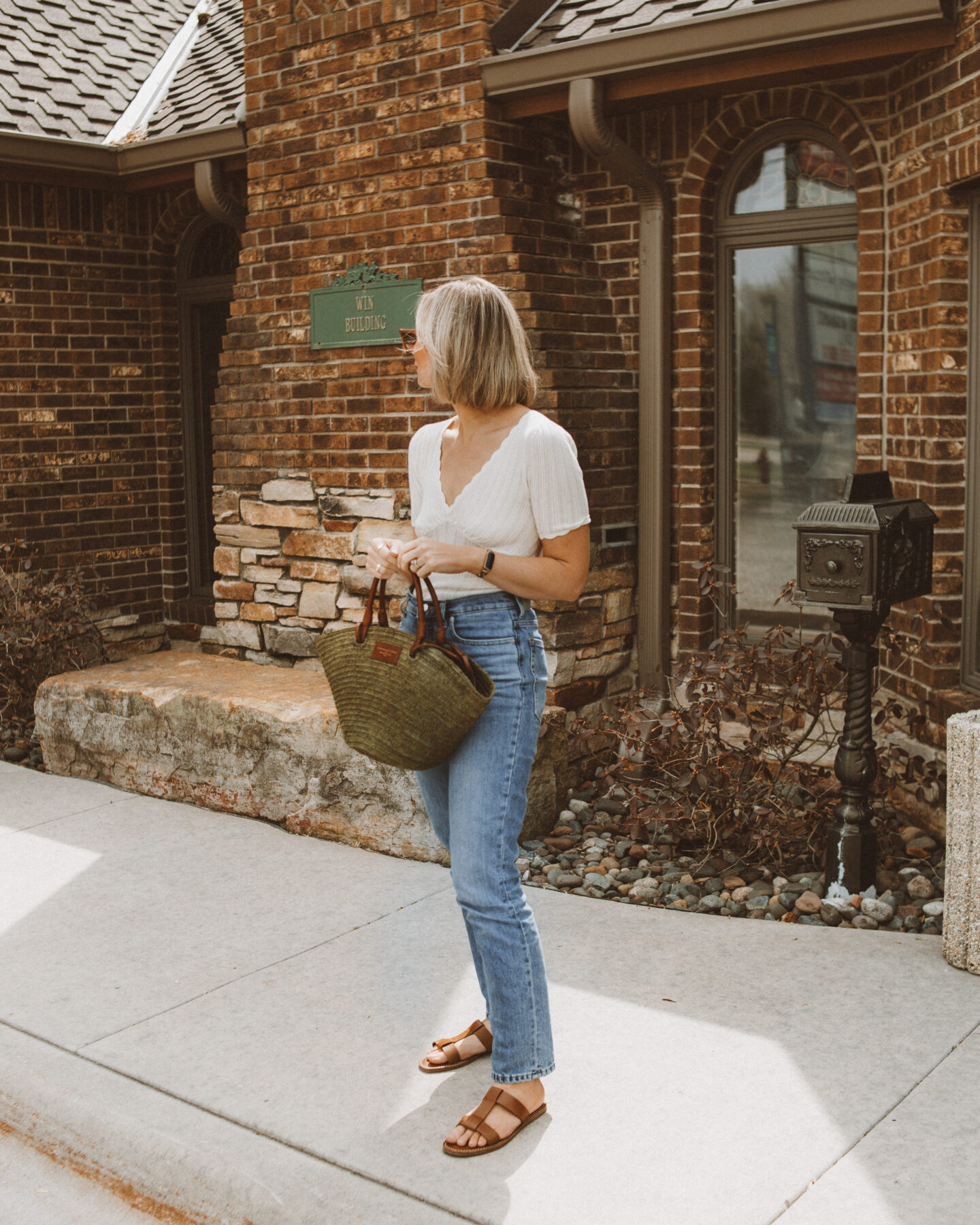 Karin Emily stands in front of a brick building wearing a white v neck tee from Sezane, a pair of blue jeans, and a khaki green justine basket bag