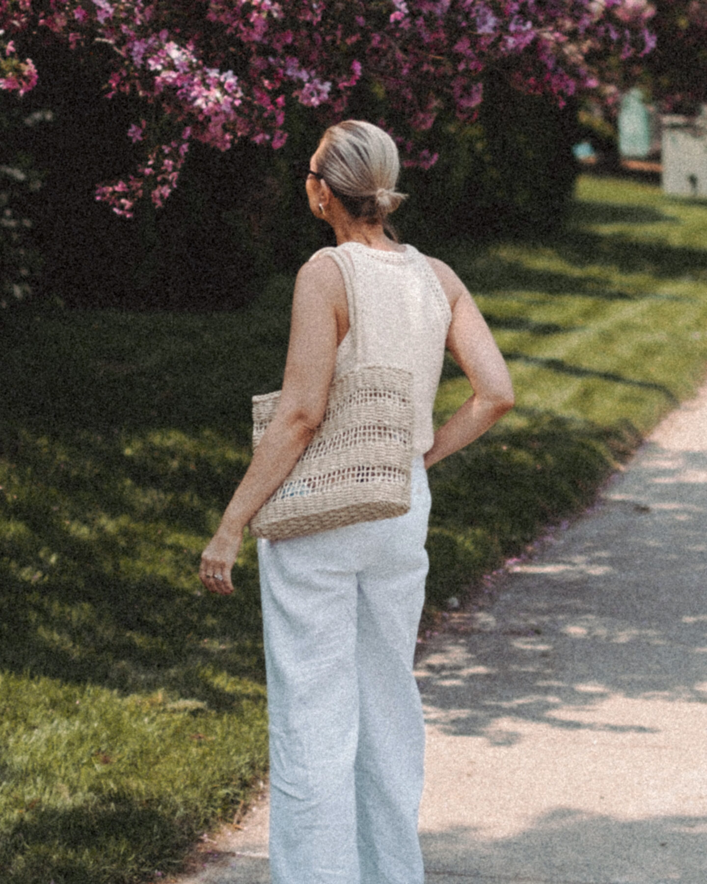 Karin Emily wears a cream sweater tank and white linen pair of pants while walking in front of a row of lilac bushes