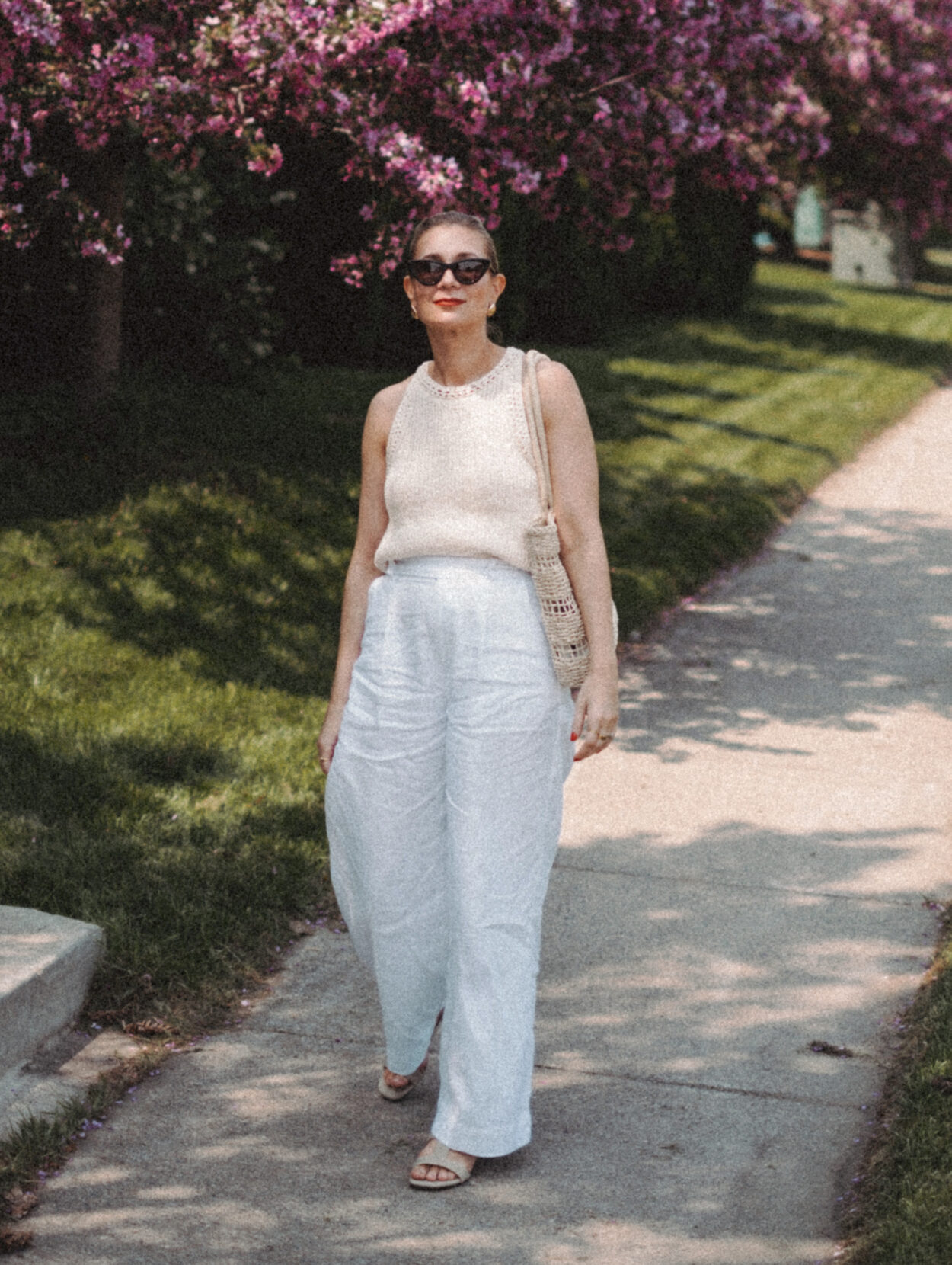 Karin Emily wears a cream sweater tank and white linen pair of pants while walking in front of a row of lilac bushes