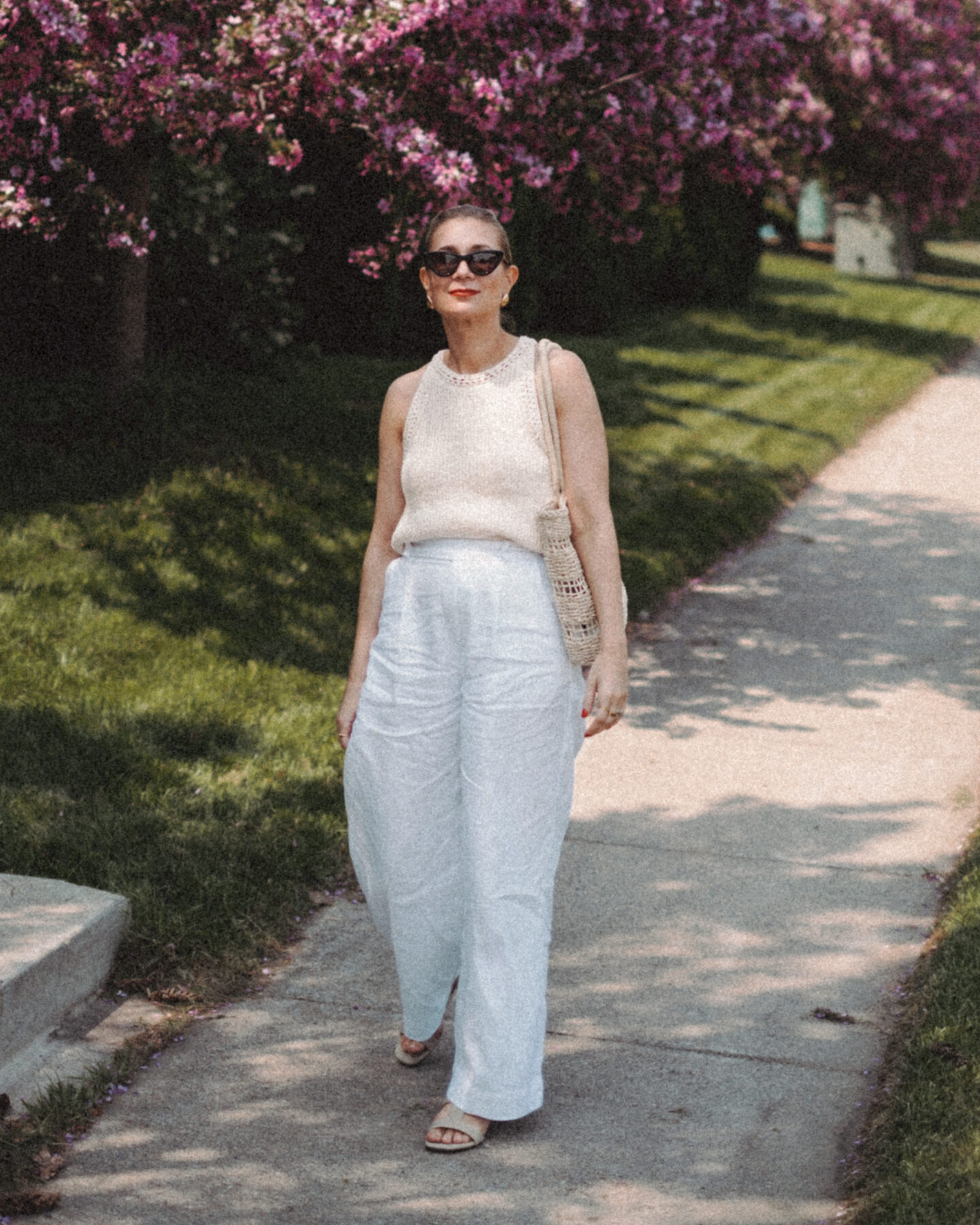 Karin Emily wears a cream sweater tank and white linen pair of pants while walking in front of a row of lilac bushes 