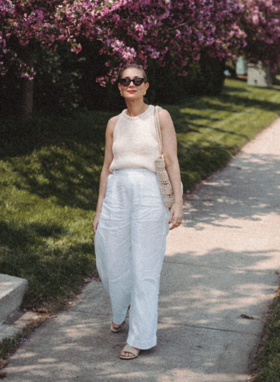 Karin Emily wears a cream sweater tank and white linen pair of pants while walking in front of a row of lilac bushes