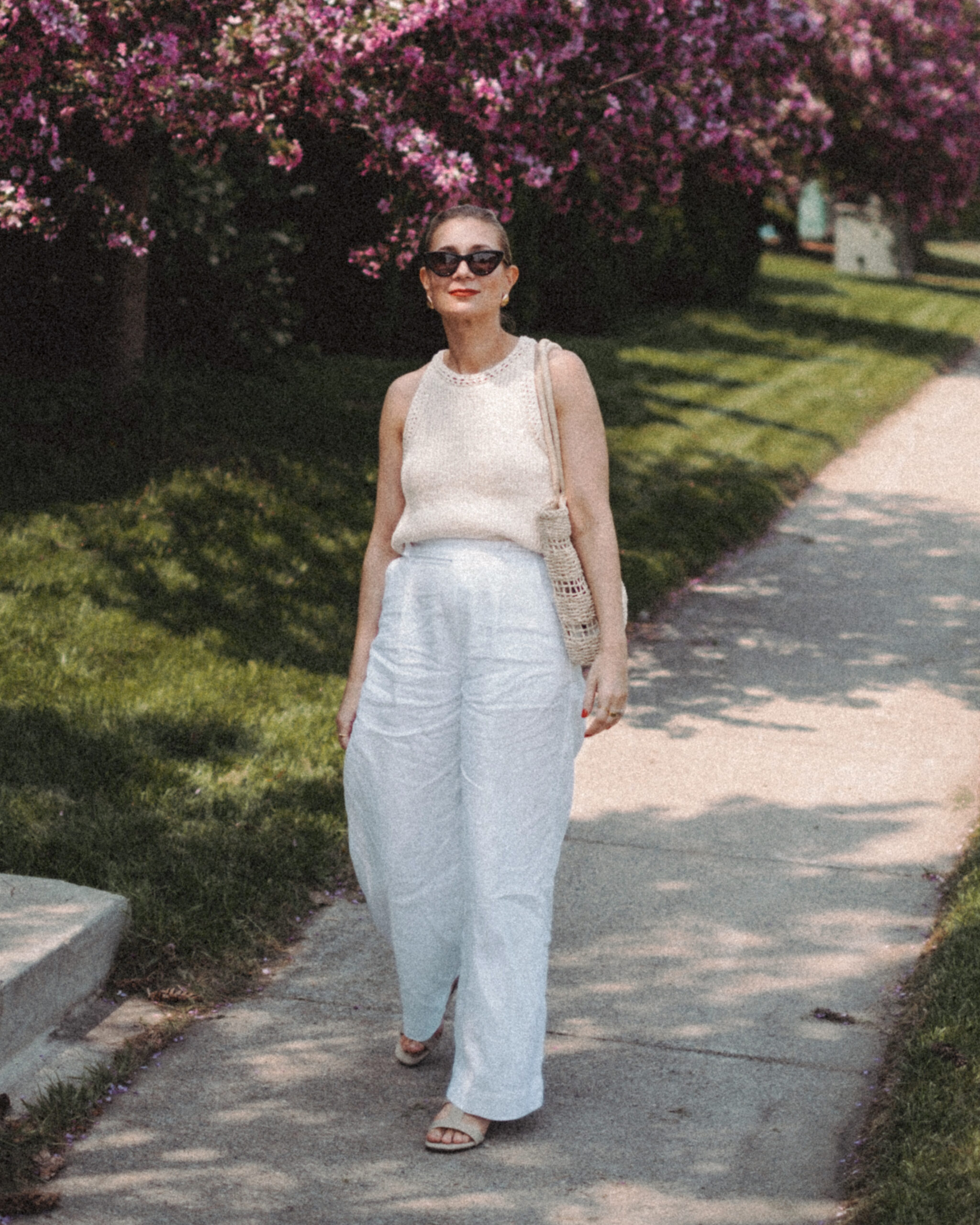 Karin Emily wears a cream sweater tank and white linen pair of pants while walking in front of a row of lilac bushes