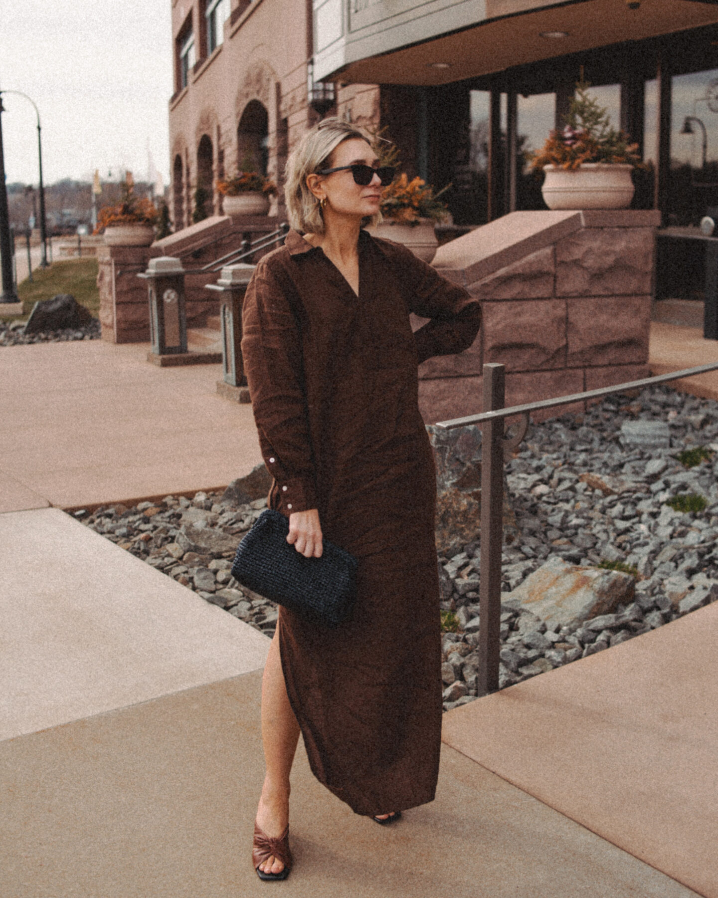 Karin Emily wears a brown linen maxi dress and brown knot sandal heels in front of a brown rock building