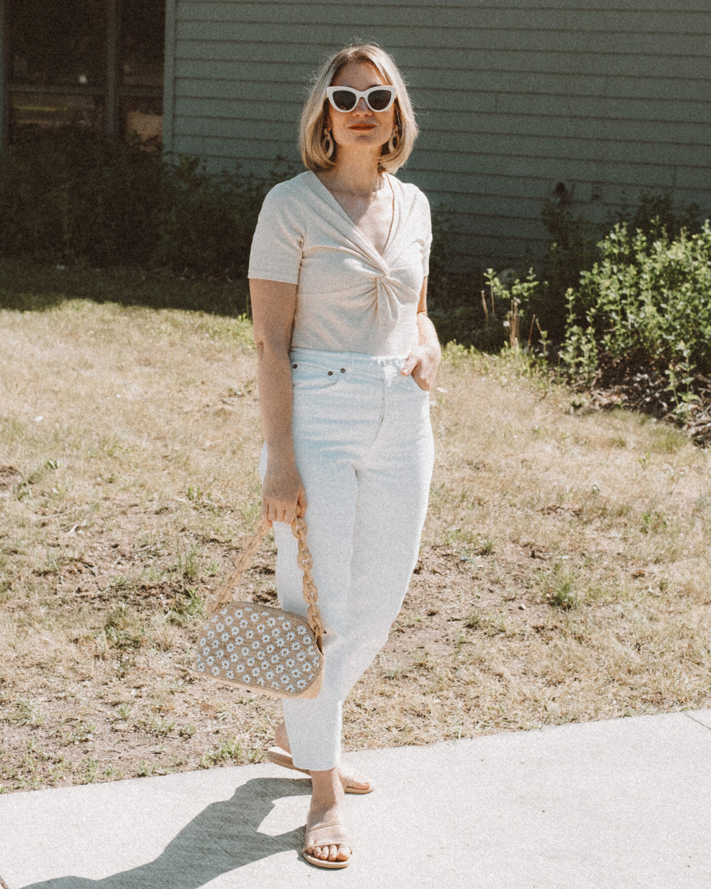 Karin Emily holds a basket bag embroidered with daisies from Sezane while wearing a knitted t-shirt and standing in front of a navy blue building