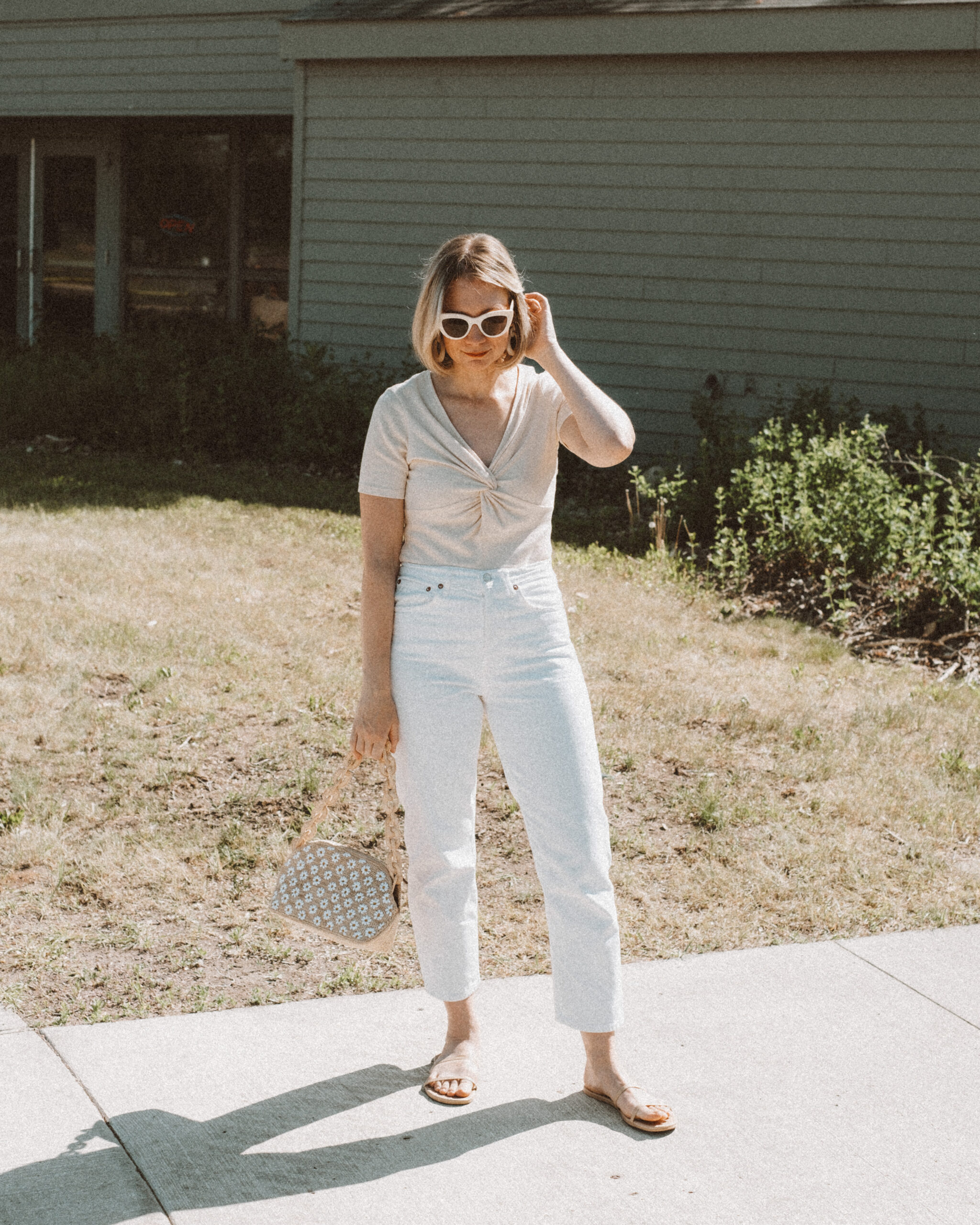 Karin Emily holds a basket bag embroidered with daisies from Sezane while wearing a knitted t-shirt and standing in front of a navy blue building