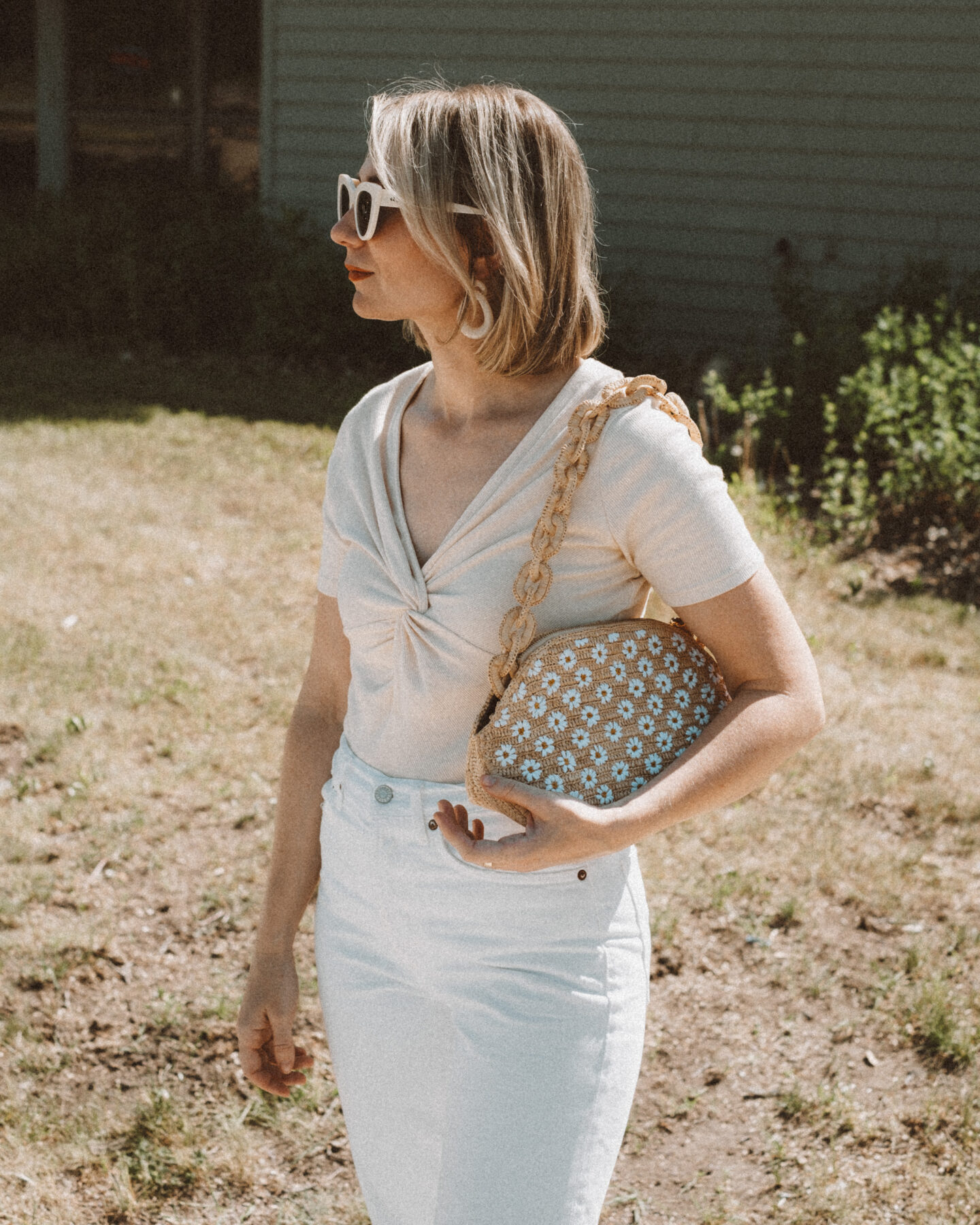 Karin Emily holds a basket bag embroidered with daisies from Sezane while wearing a knitted t-shirt and standing in front of a navy blue building 