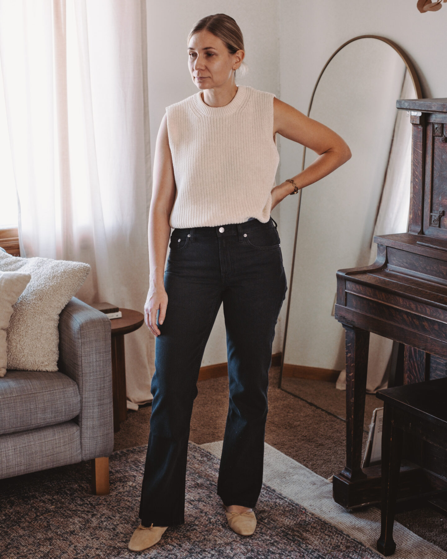 Karin Emily stands in her living room wearing a pair of bootcut jeans and a sweater tank