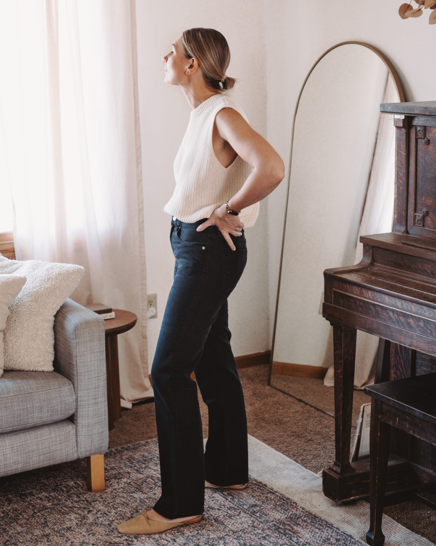 Karin Emily stands in her living room wearing a pair of bootcut jeans and a sweater tank