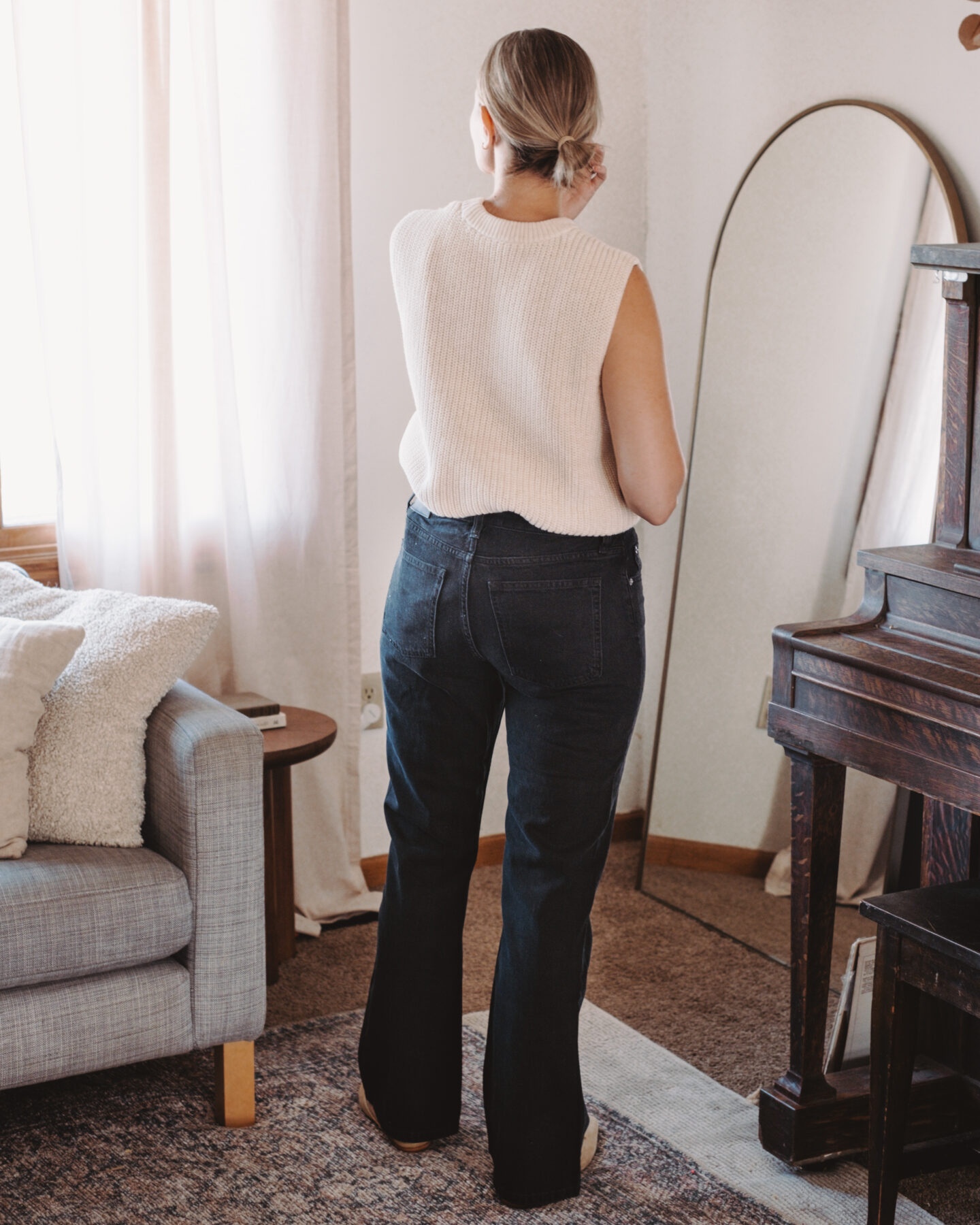 Karin Emily stands in her living room wearing a pair of bootcut jeans and a sweater tank