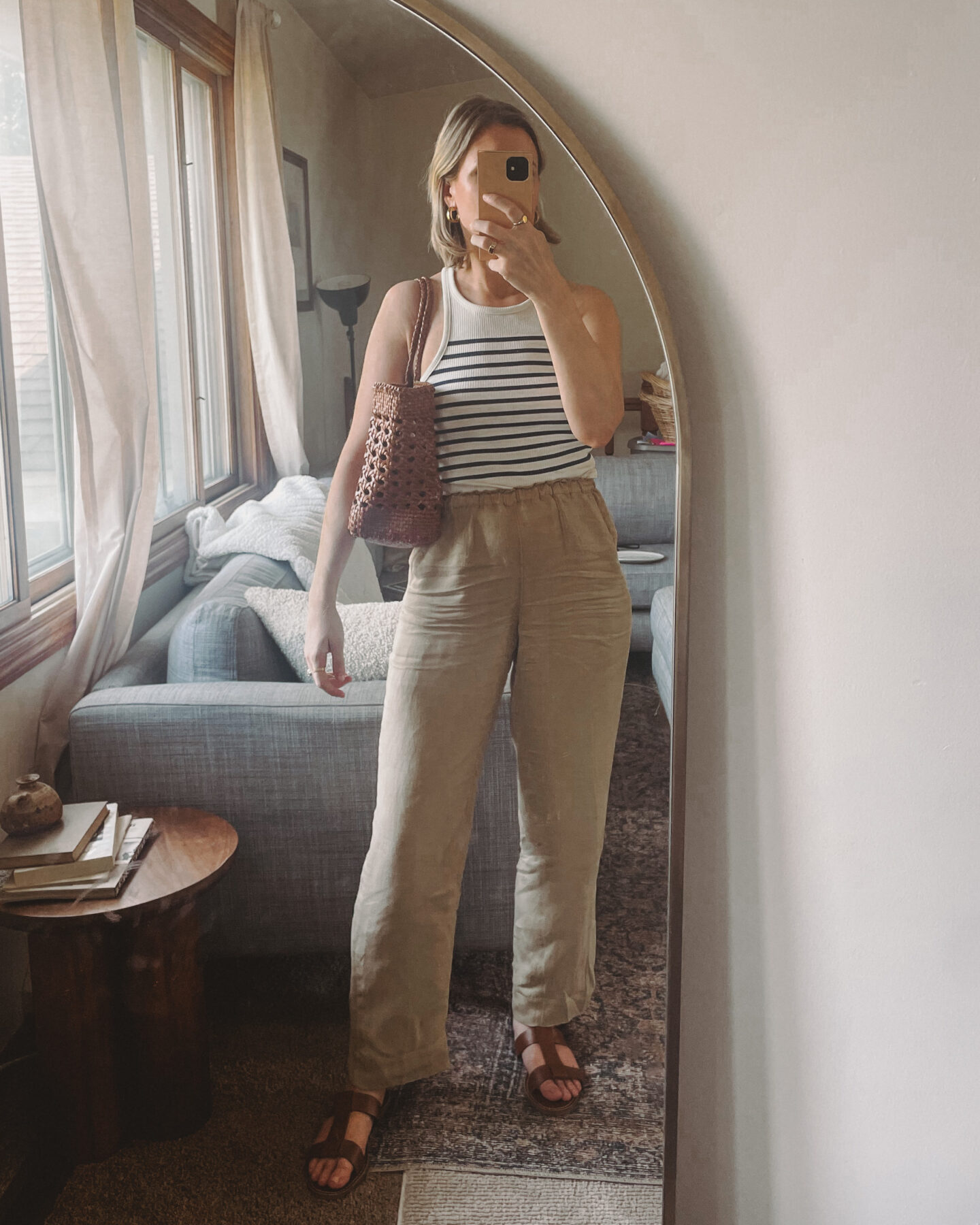 Karin Emily wears a striped tank, brown linen pants, and brown accessories while taking a mirror selfie in her living room
