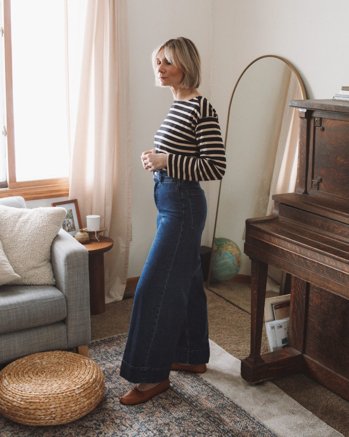 Karin Emily stands in her living room, wearing a striped long sleeve tea from Everlane and a dark wash pair of wide leg jeans