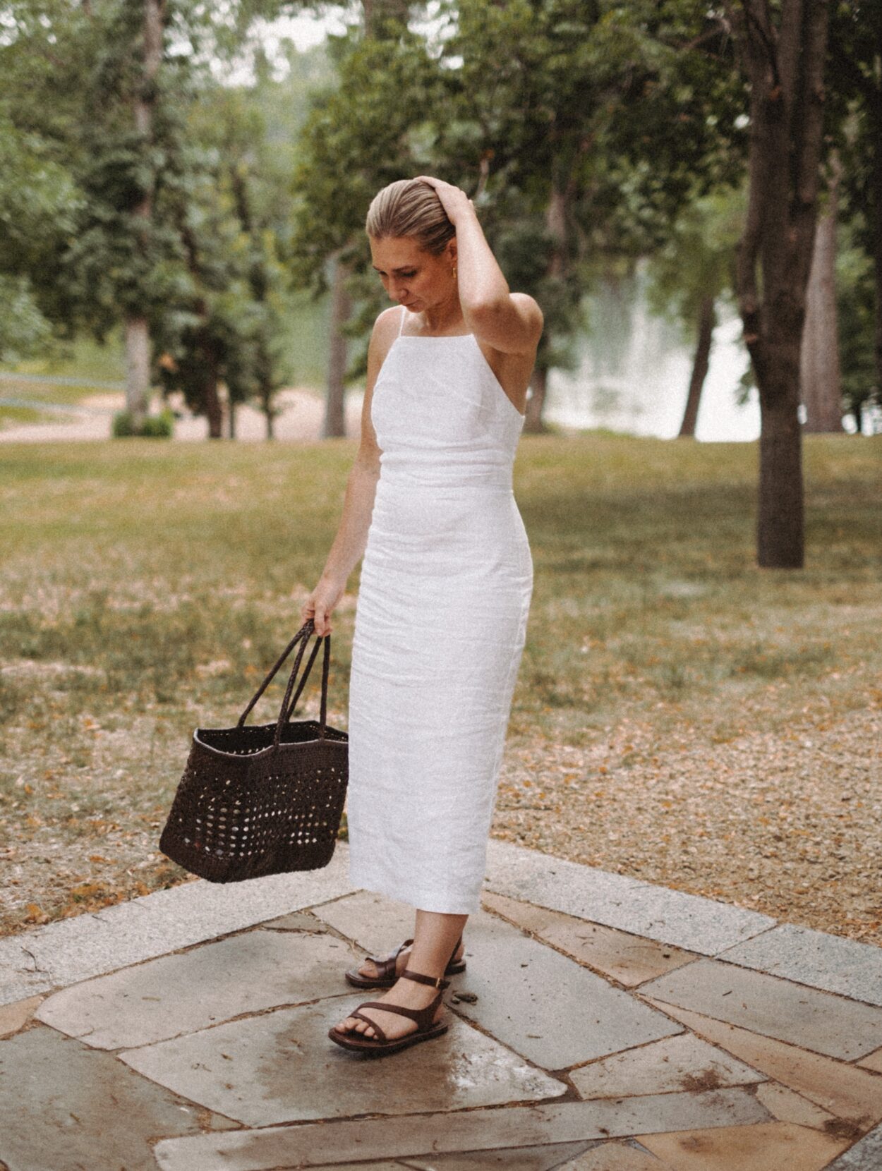 Karin Emily stands in front of a lake wearing a white linen dress, brown woven bag, and dark brown sandals from Madewell