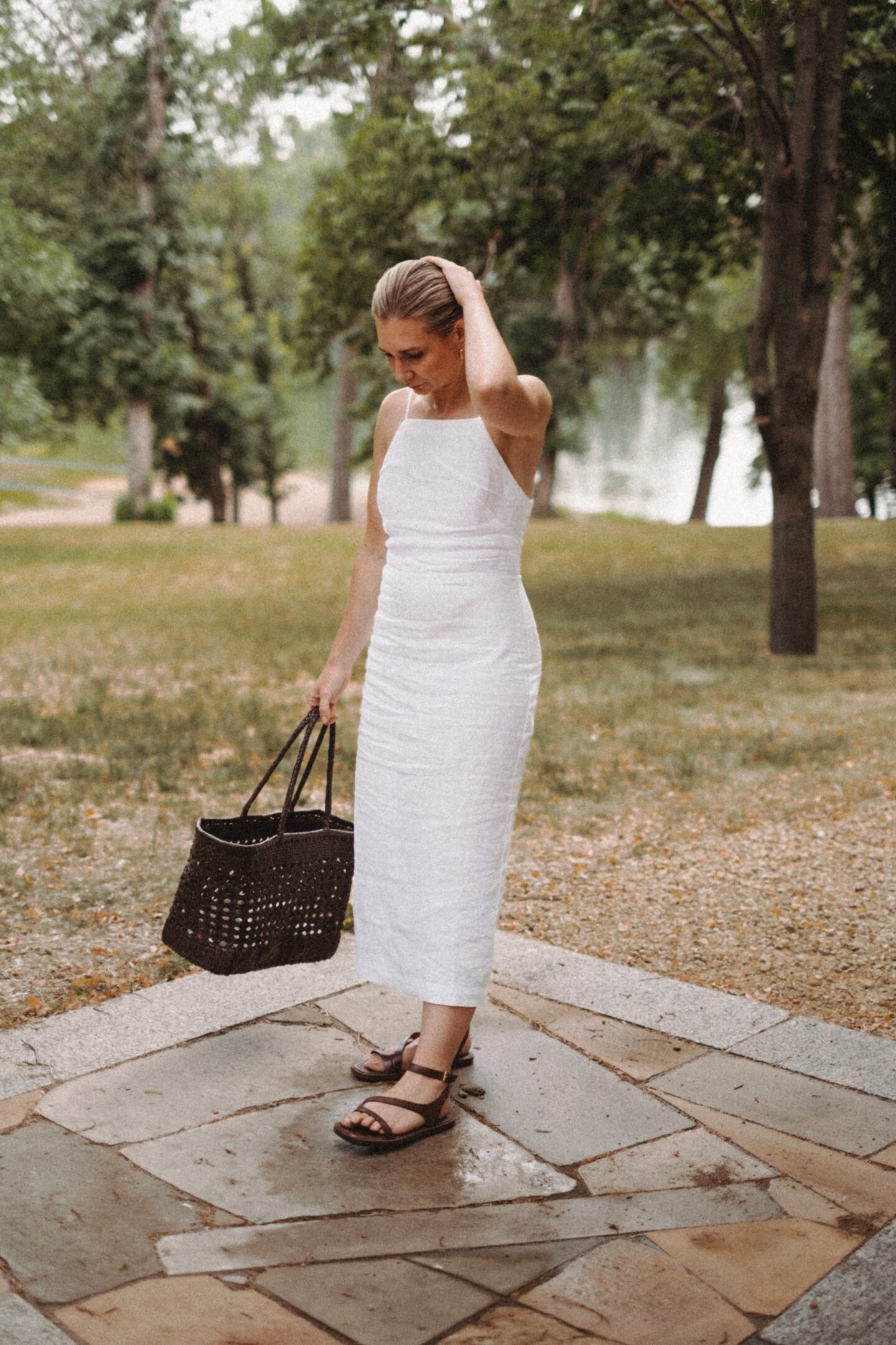 Karin Emily stands in front of a lake wearing a white linen dress, brown woven bag, and dark brown sandals from Madewell