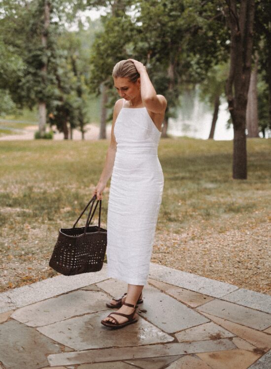 Karin Emily stands in front of a lake wearing a white linen dress, brown woven bag, and dark brown sandals from Madewell