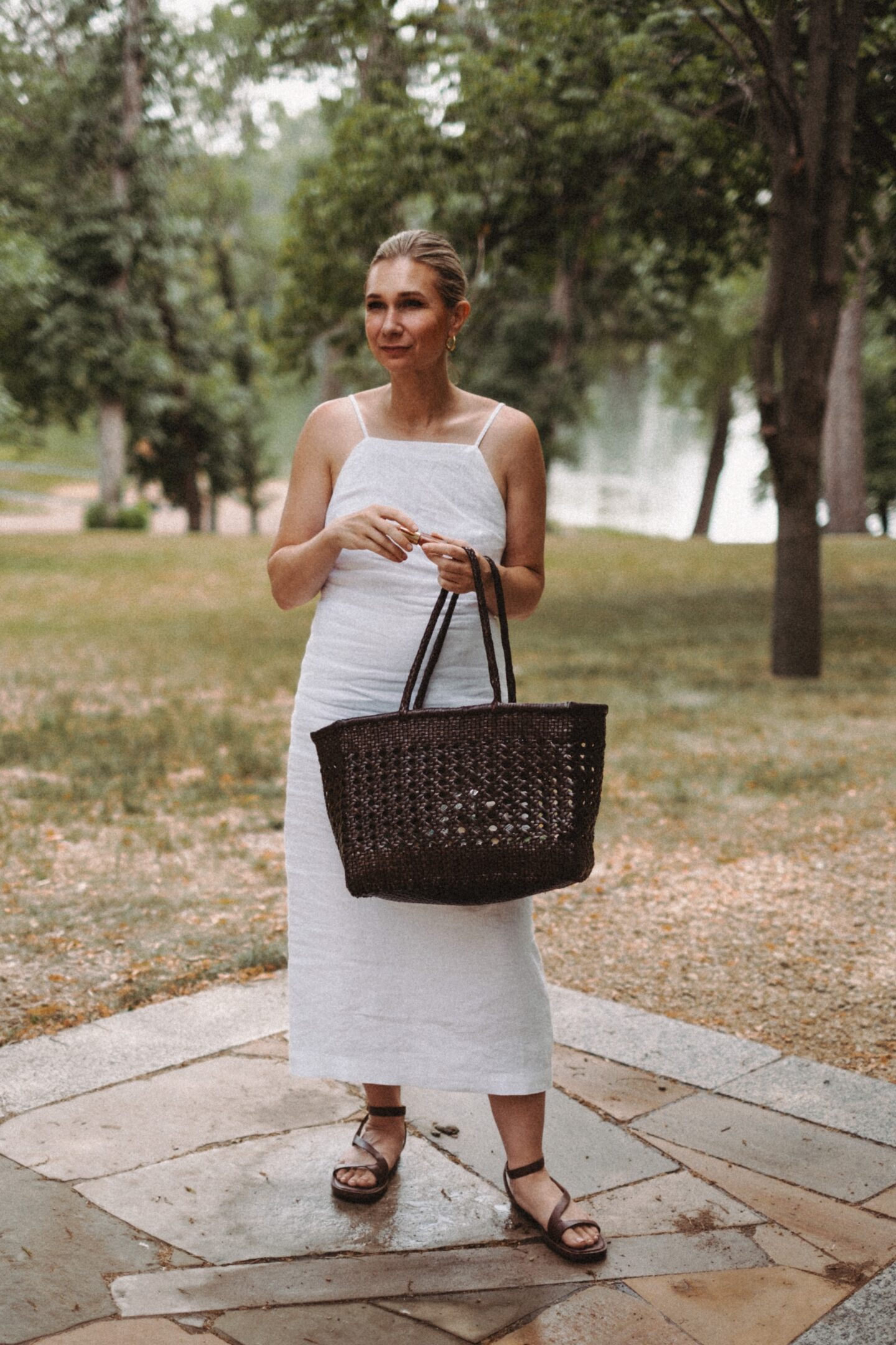 Karin Emily stands in front of a lake wearing a white linen dress, brown woven bag, and dark brown sandals from Madewell