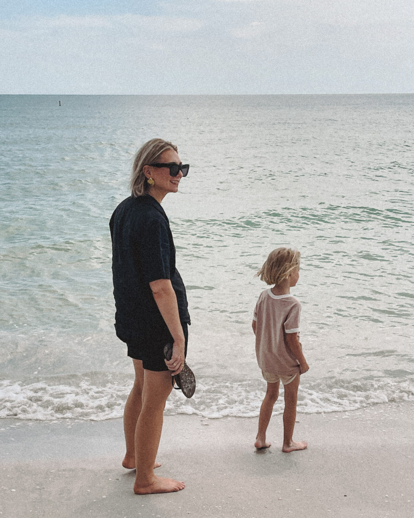 Karin Emily wearing a blacking coordinating linen set from AYR stands in the sand on the beach while looking at the water with her daughter