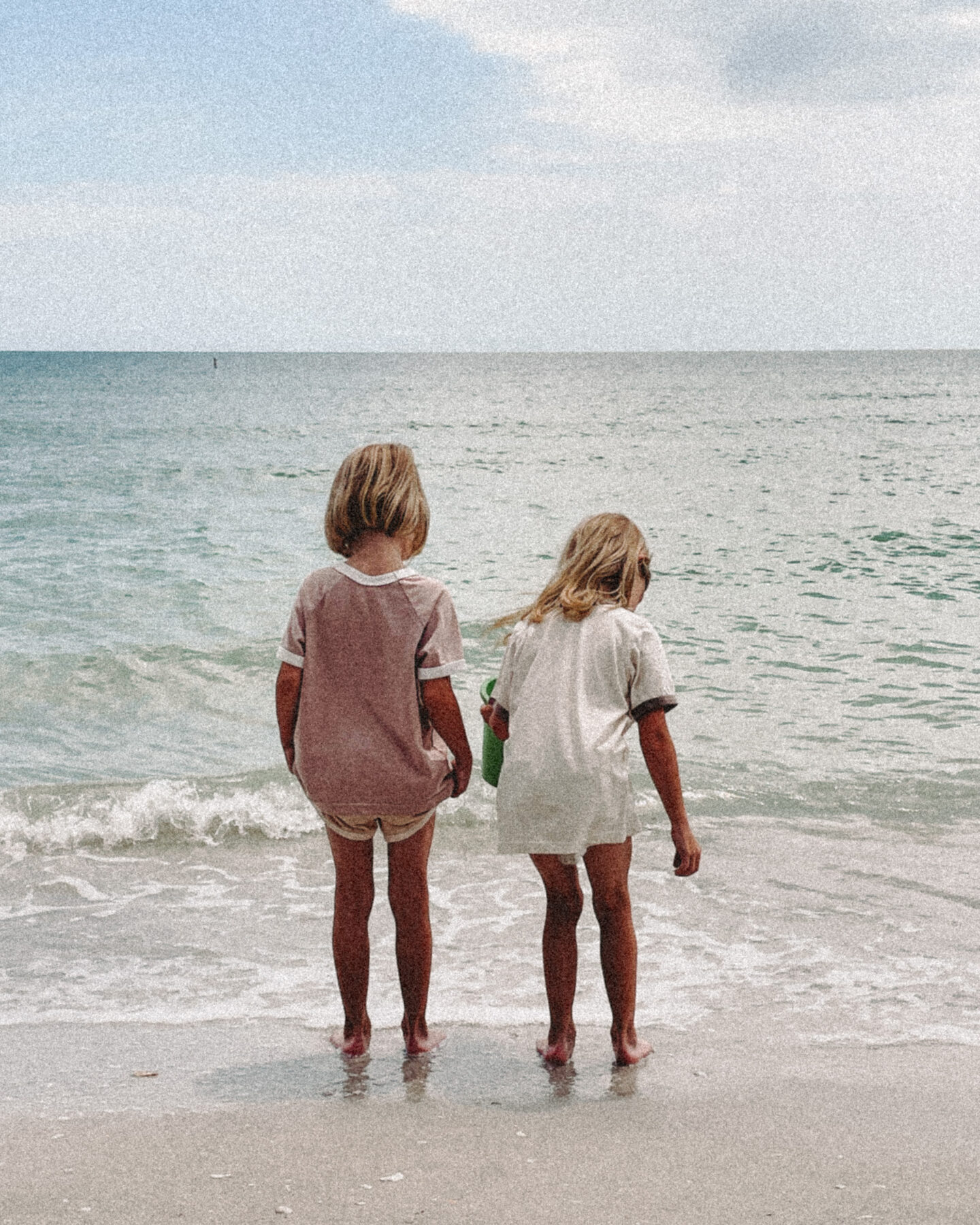 Twin girls look out at the ocean water while standing in the sand on the beach