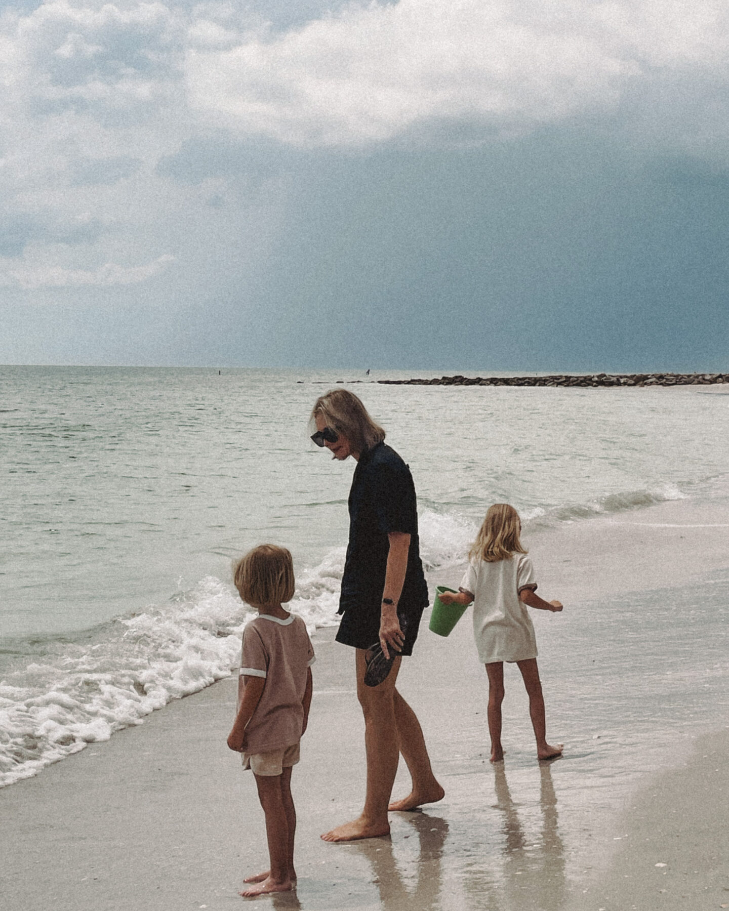 Karin Emily wearing a blacking coordinating linen set from AYR stands in the sand on the beach while looking at the water with her twin daughters 