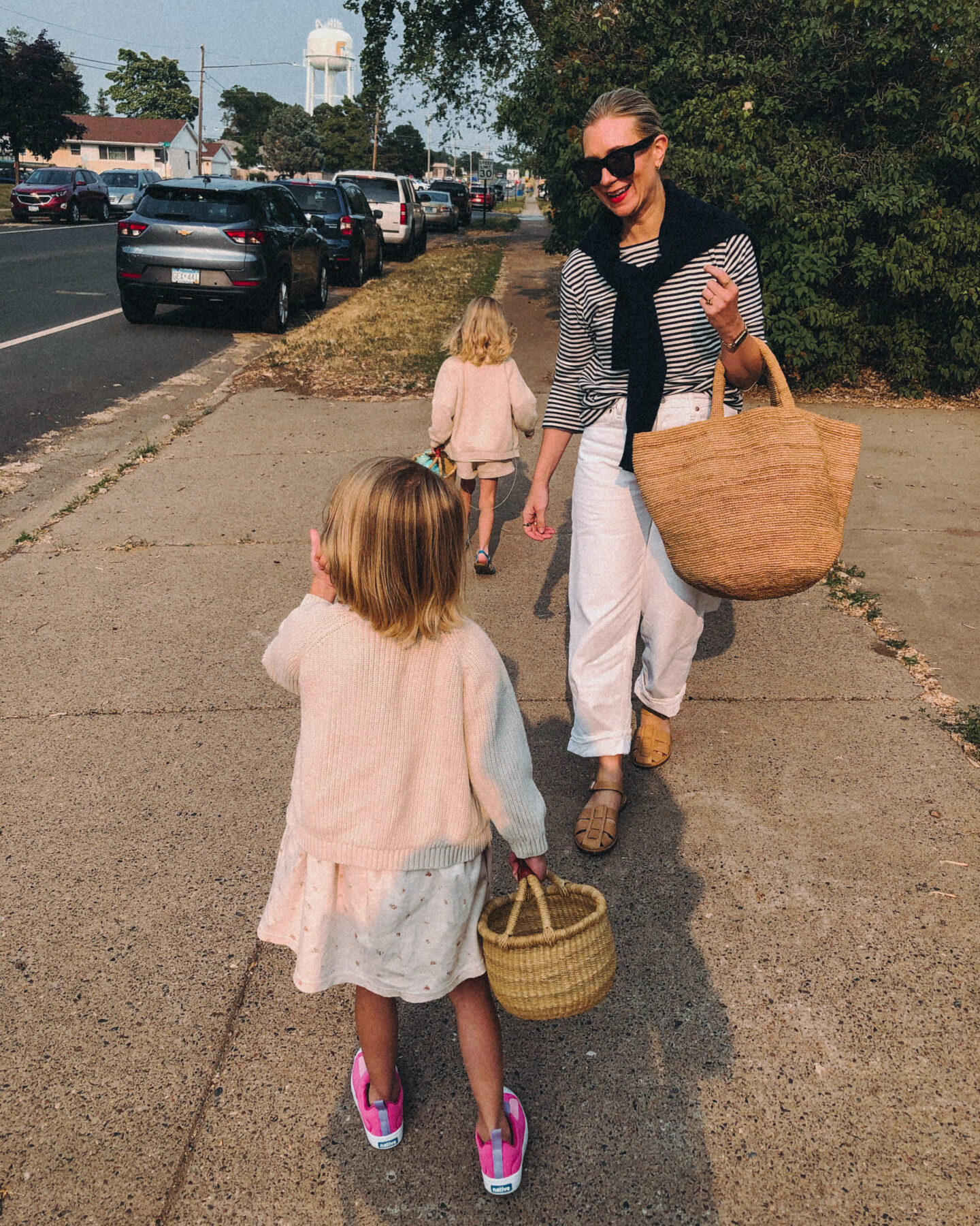 Karin Emily walks with her kids on a sidewalk and wears a black and white striped long sleeve tee white baggy jeans from agolde, nude fisherman sandals, a basket bag, and a navy cashmere sweater over her shoulders